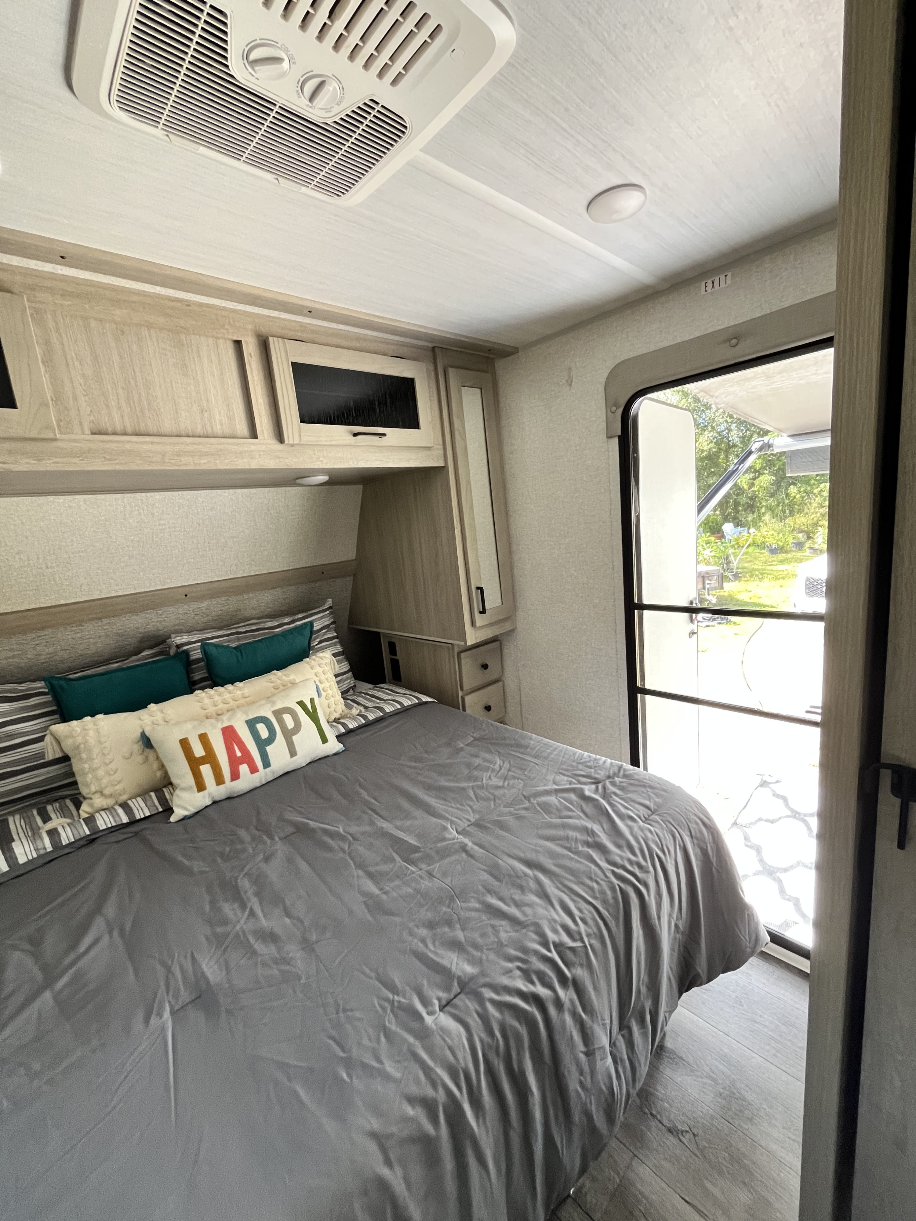 Cozy RV rental bedroom featuring a comfy bed with a "HAPPY" pillow, overhead cabinets, and an open door to nature.. East to West Alta 2022