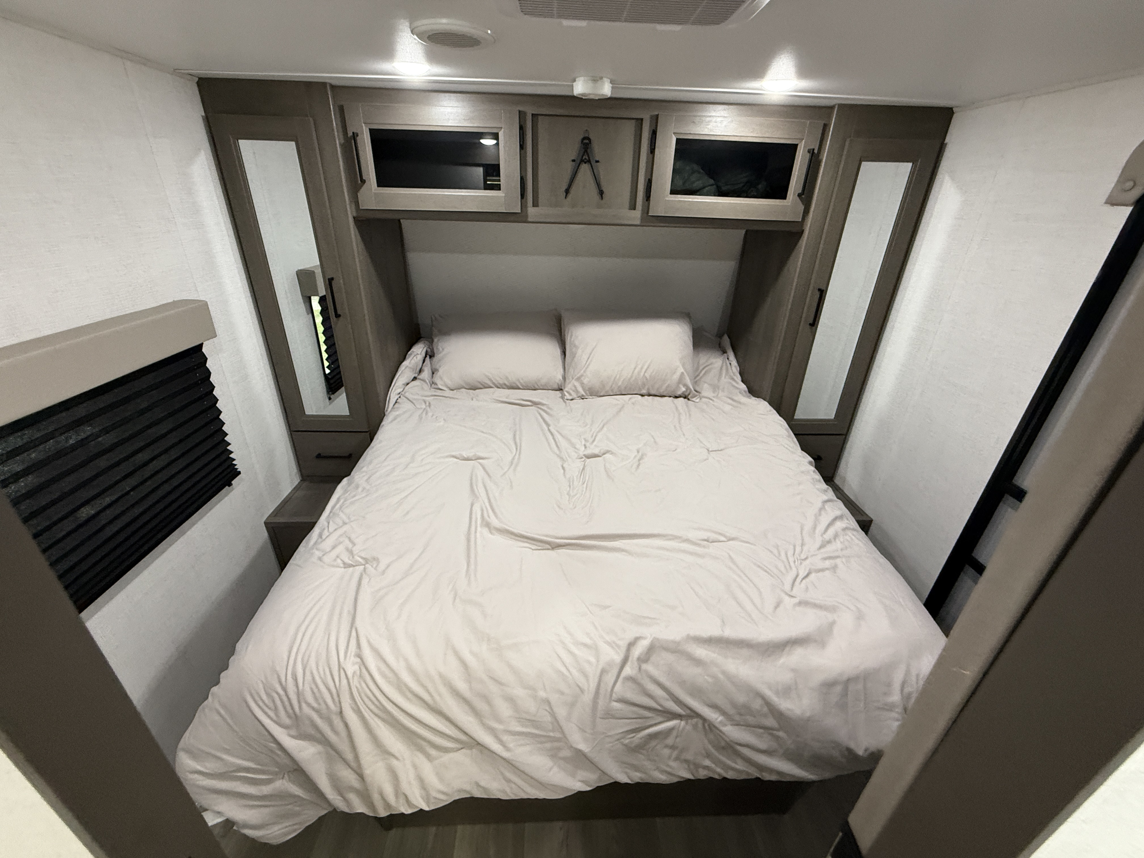 This RV rental bedroom features a bed with light-colored bedding, two pillows, overhead storage cabinets, and a side window with black blinds.. Grand Design 265-BHT 2025