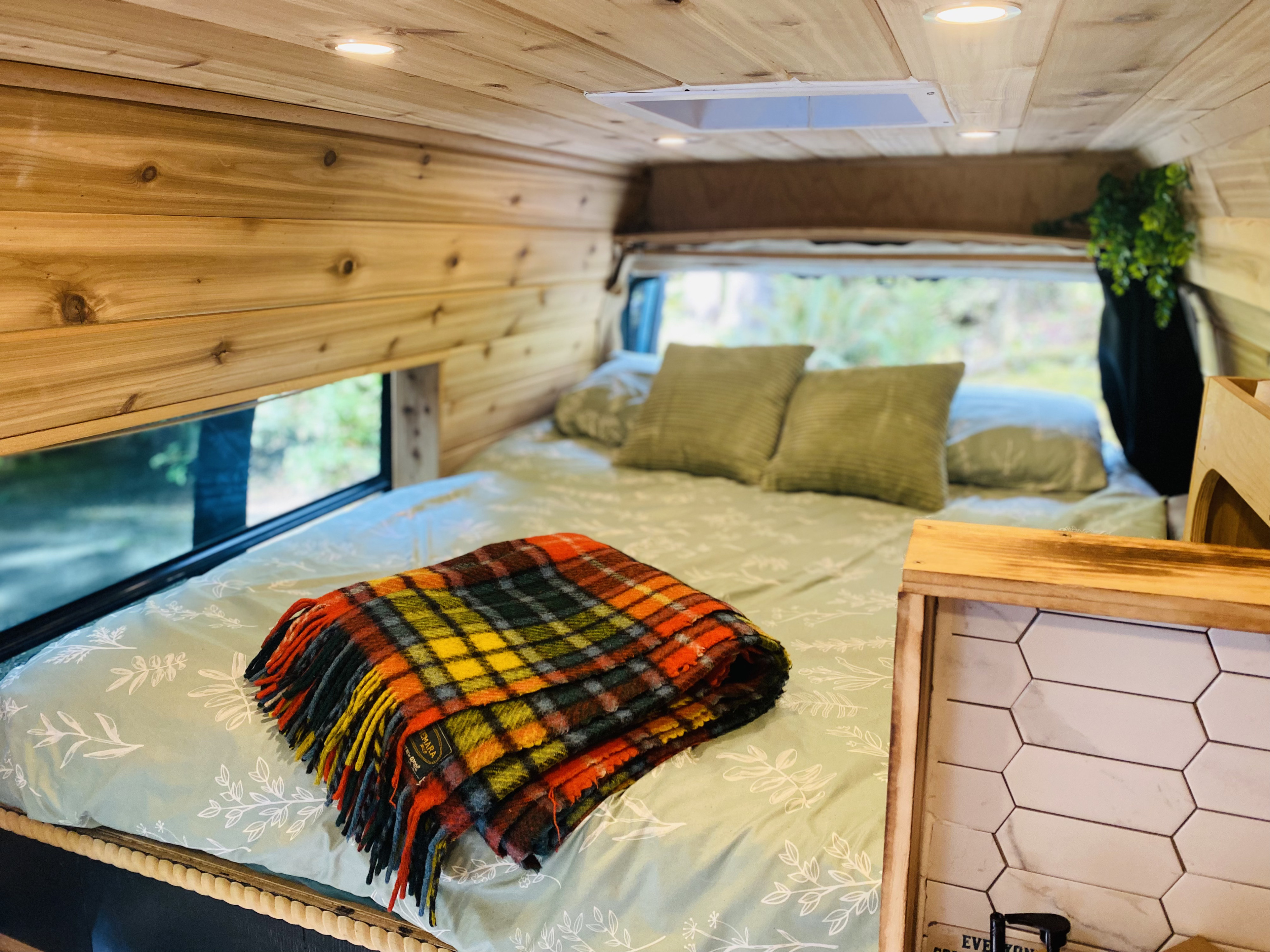 Experience comfort in this RV rental: wooden walls, cozy bed with plaid blanket, green pillows, and a peaceful window view.. Ford Econolline 250s 2001