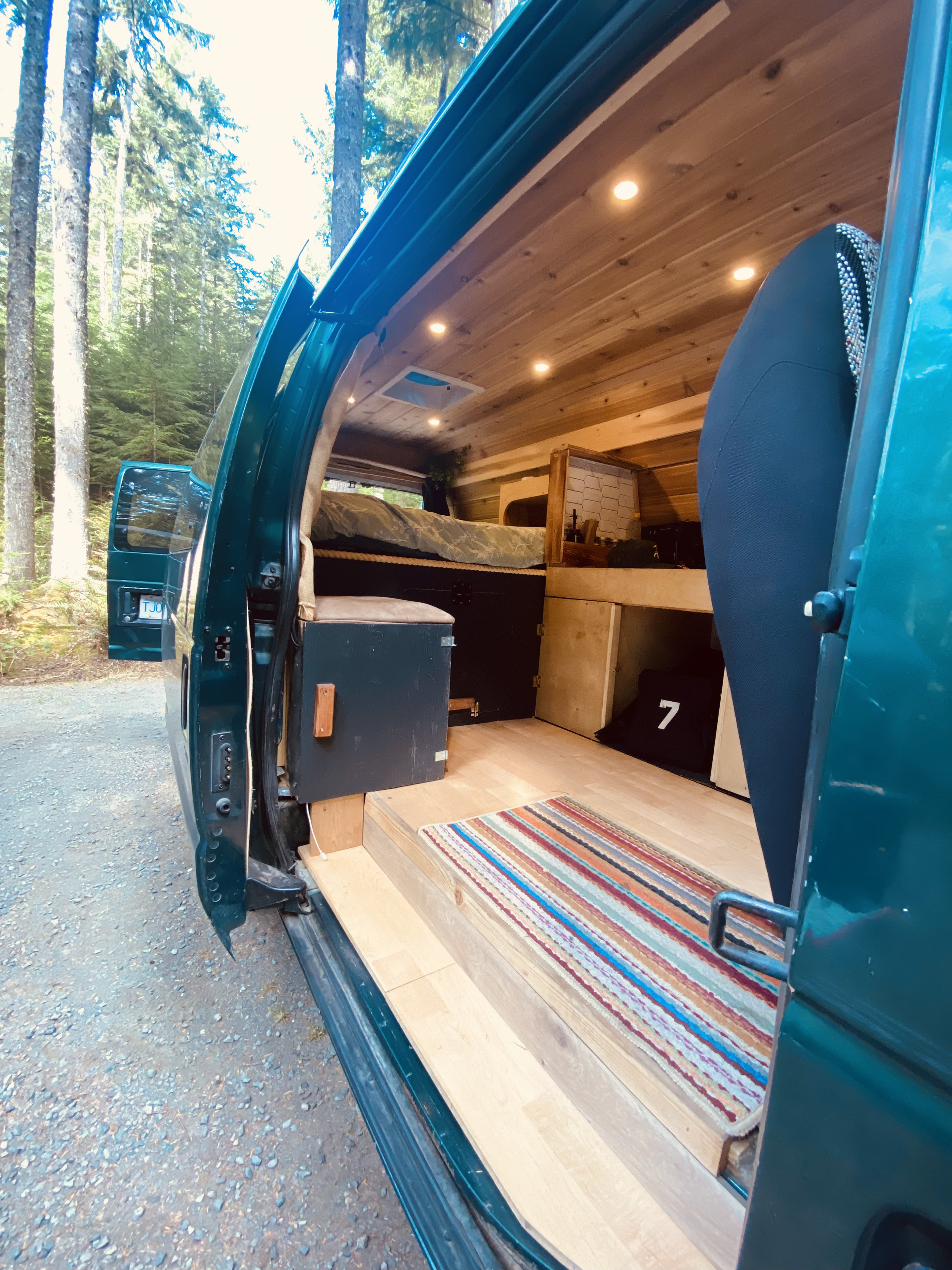 Converted RV rental van interior: a cozy wood-paneled space with bed, storage, and rug, amidst tall forest trees.. Ford Econolline 250s 2001