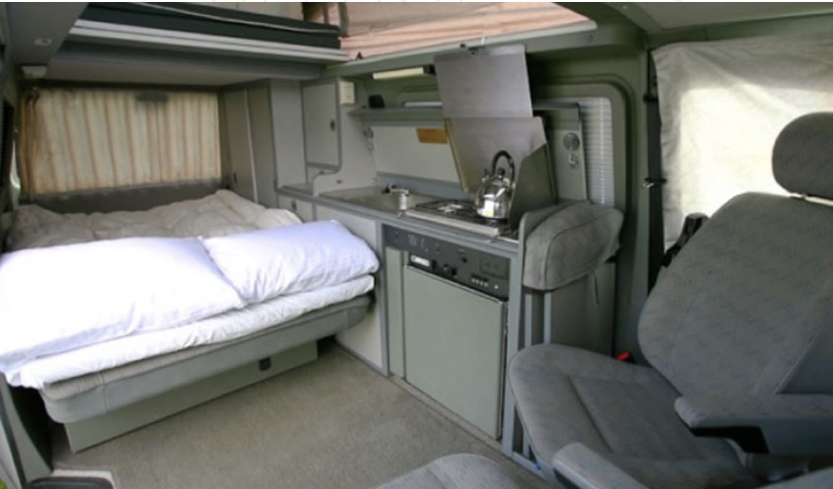 Cozy RV rental interior with bed, kitchenette (stovetop, fridge, sink), and front seat. Perfect for travel.. Volkswagen Multivan 2001