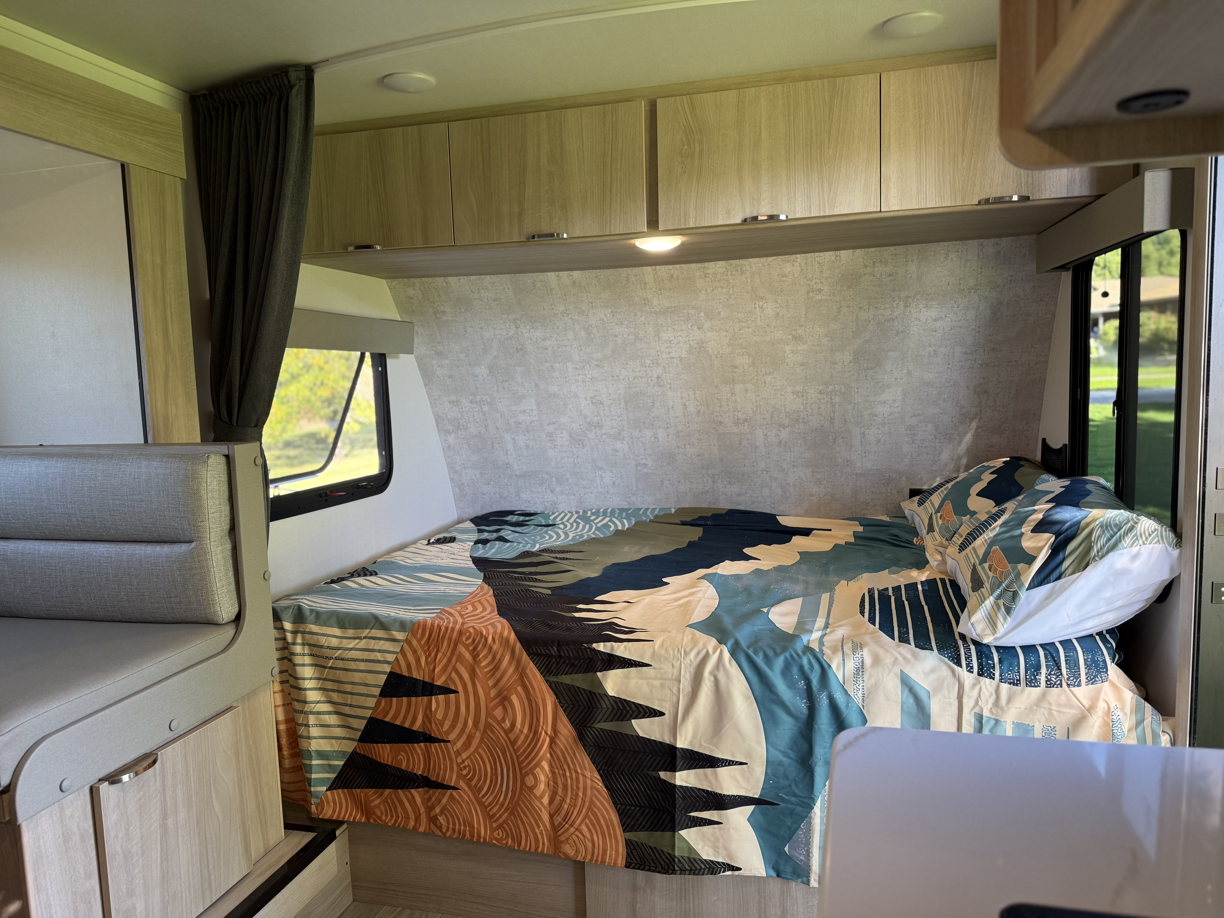 Modern RV rental interior: a cozy bedroom with a colorful bedspread, overhead cabinets, window, and dinette seating.. Winnebago Micro Minnie 2100BH 2025