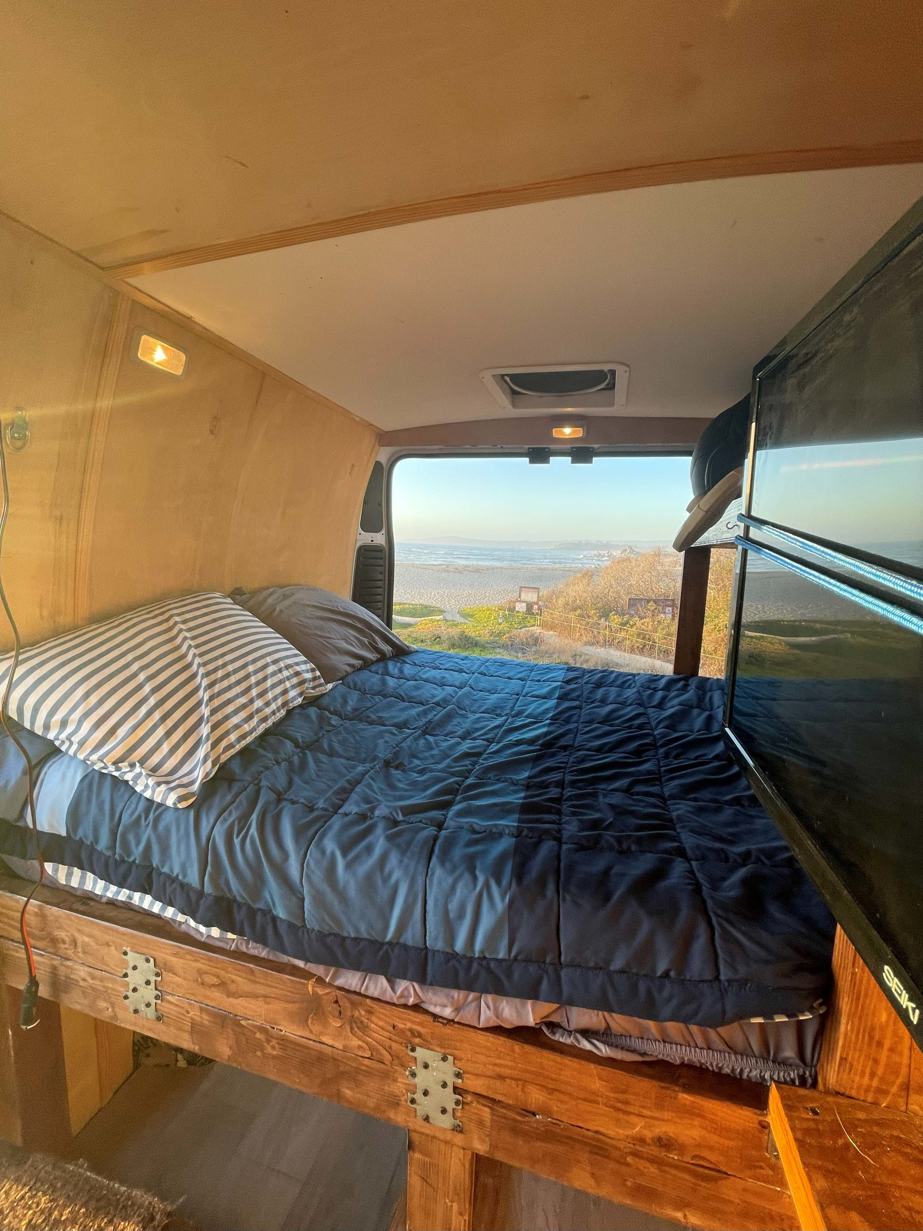 Inside a comfy RV rental: The bed with blue comforter and striped pillow overlooks a beautiful beach and ocean view, with a TV.. Dodge 1500 2019