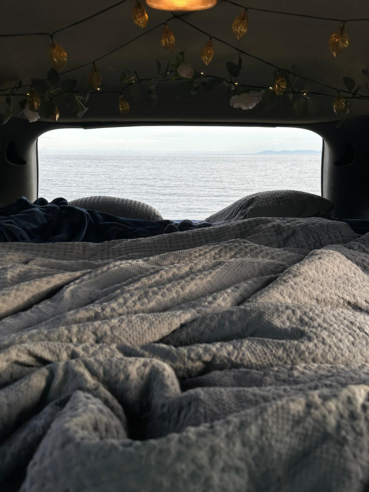 View from inside an RV rental: a cozy bed with gray blankets, decorated string lights, and a serene ocean vista.. Dodge Caravan 2005