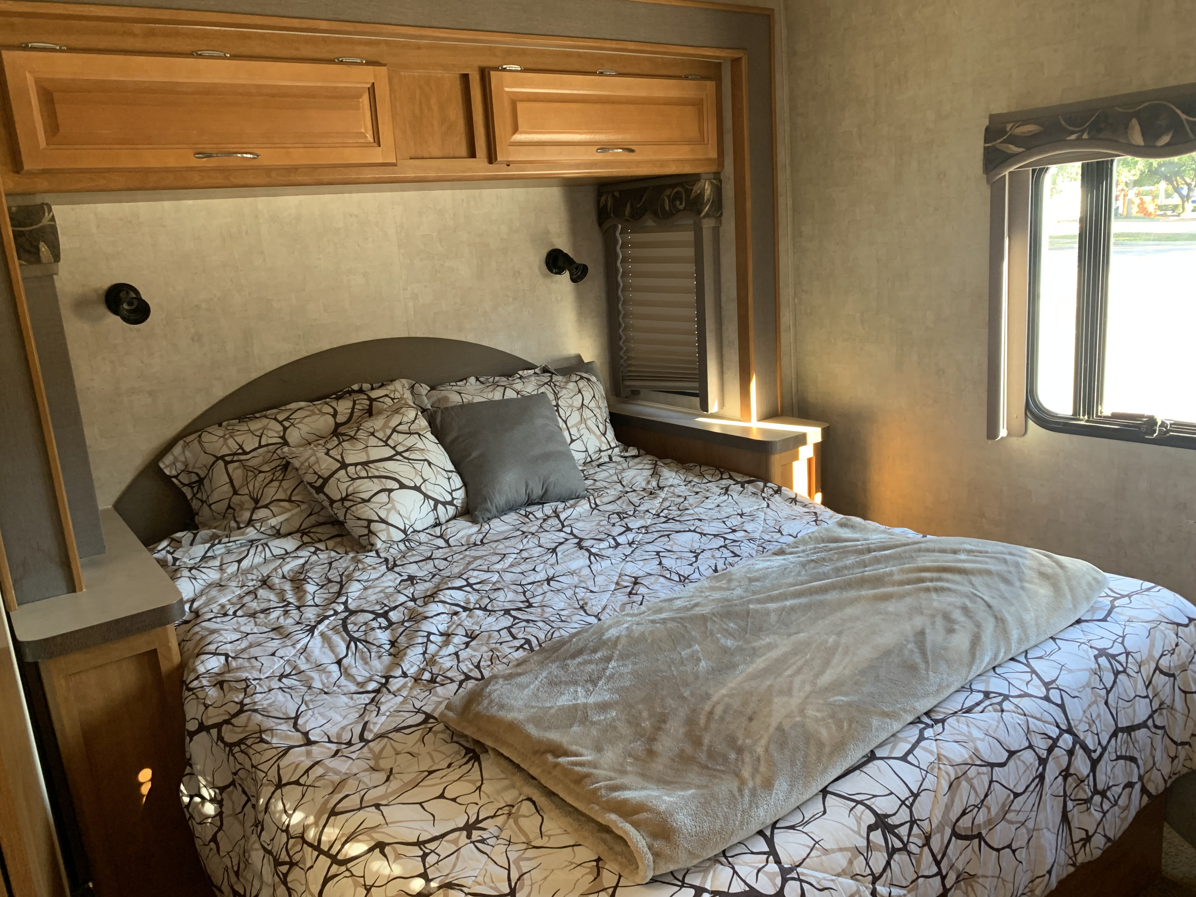Experience comfort in this RV rental bedroom with branch-patterned bedding, a cozy blanket, overhead cabinets, and a bright window.. Jamboree Jamboree Motorhome 2006