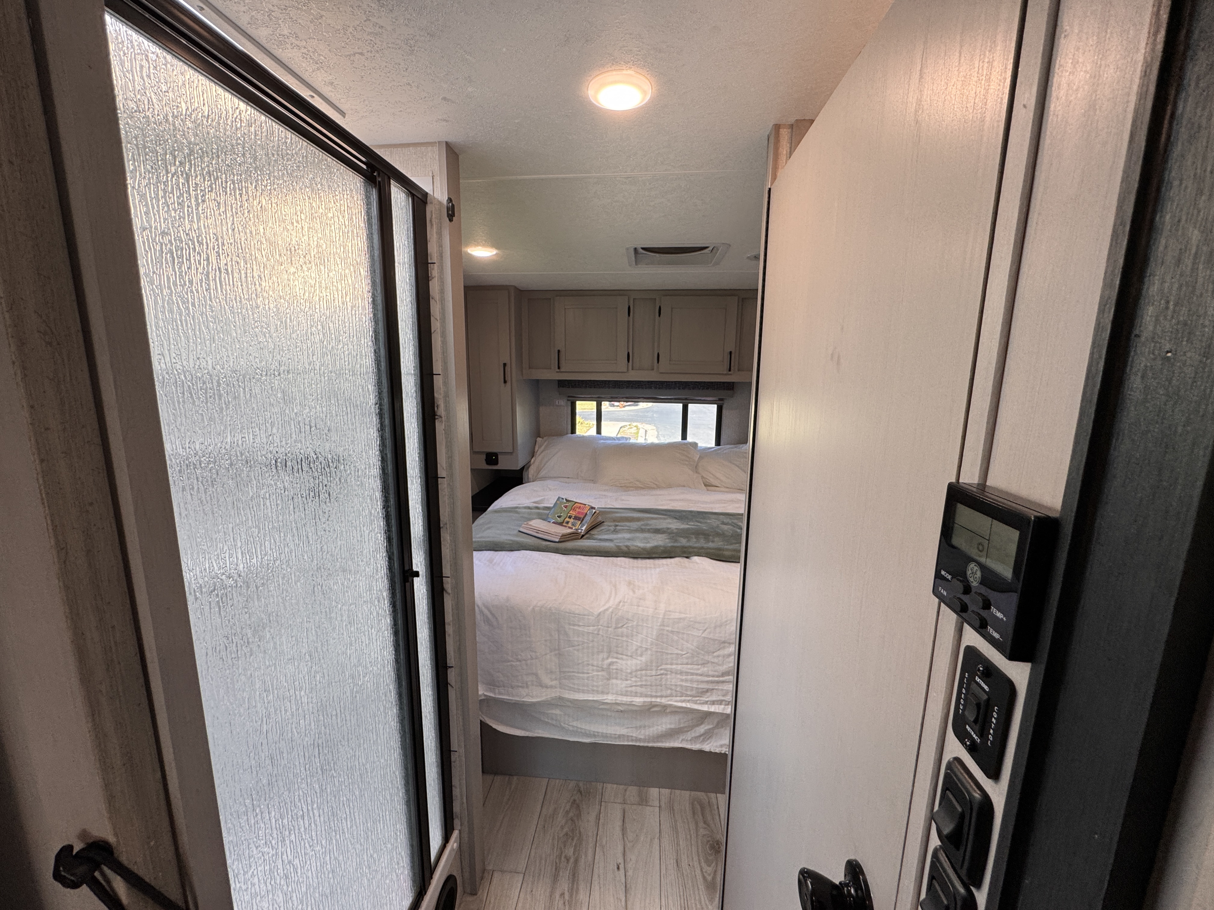 RV rental interior: A narrow hallway reveals a cozy bedroom with a bed, cabinets, and window. A textured glass shower is on the left.. Forest River Coachmen Freelander 2025