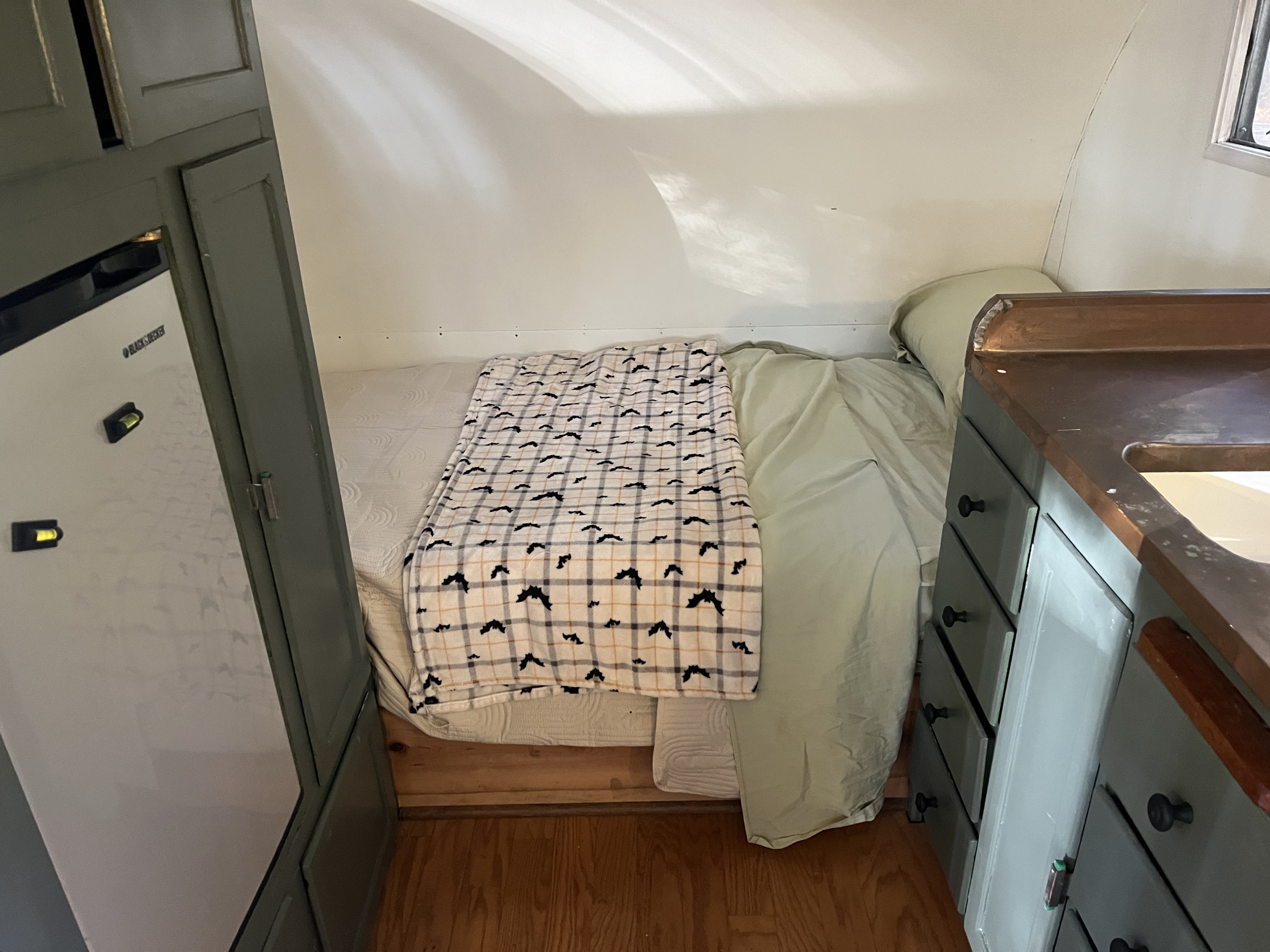 Cozy RV rental interior with a bed, bat-patterned blanket, compact fridge, and a small kitchenette with wooden counters.. Shasta 1500 1954