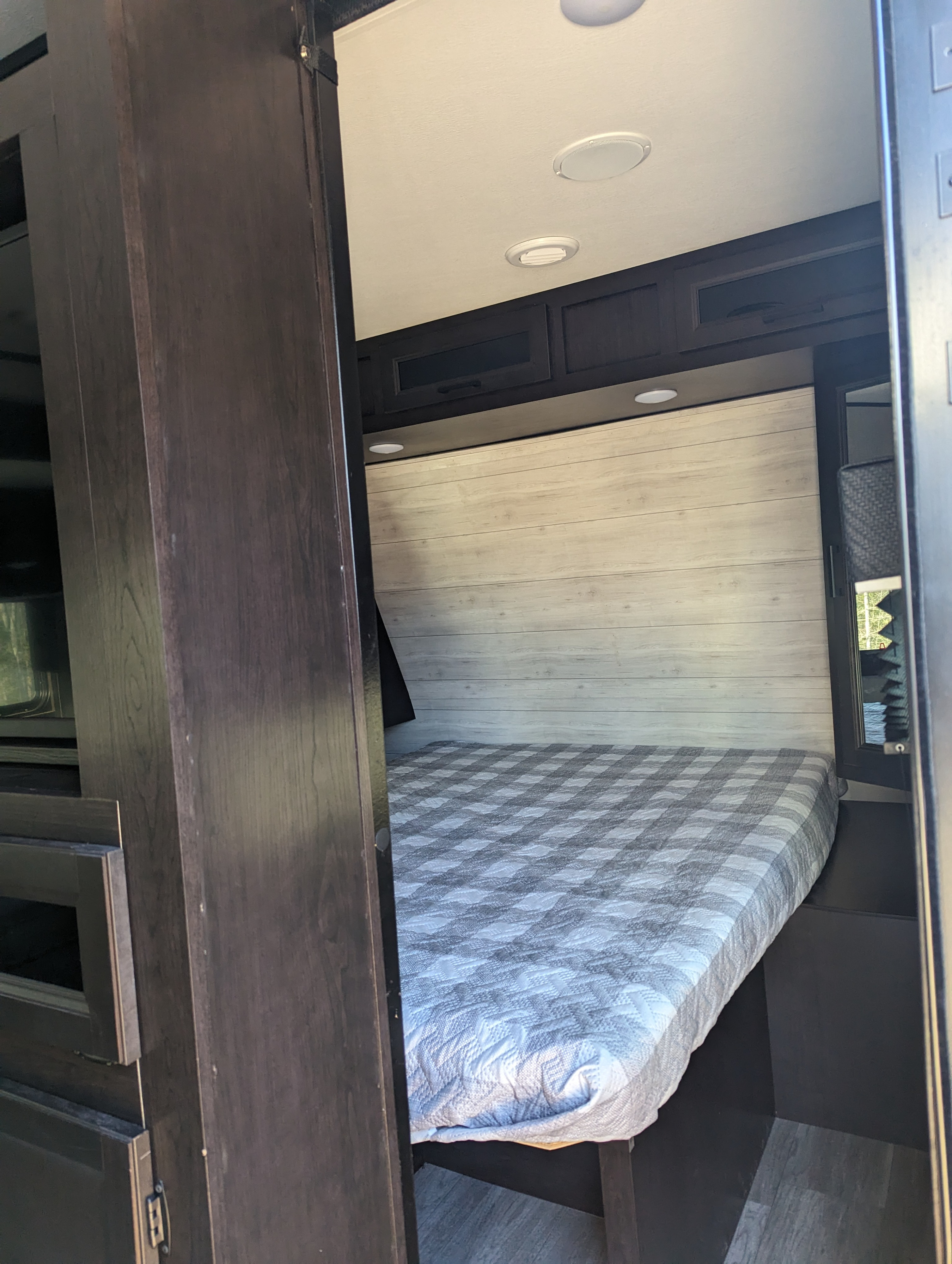 View of a modern rv rental bedroom: a cozy bed with a grey plaid comforter, light wood headboard, dark cabinets & ceiling lights.. Jayco Jay Flight 2022