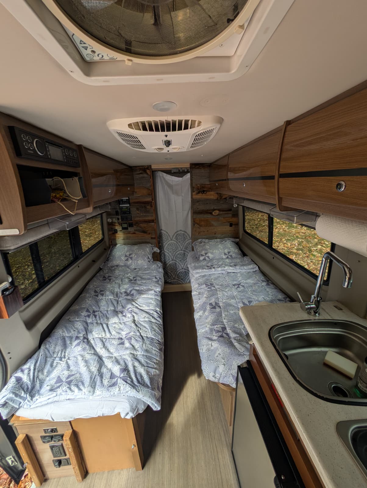 Inside this cozy rv rental: twin beds with patterned comforters, a compact kitchen with sink, wood walls, and overhead storage.. Winnebago Travato 2016