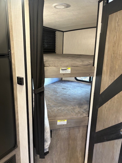 RV rental bunk bed area: two stacked beds with mattresses, partially enclosed by a black curtain, within a compact camper interior.. Heartland RVs North Trail 2021