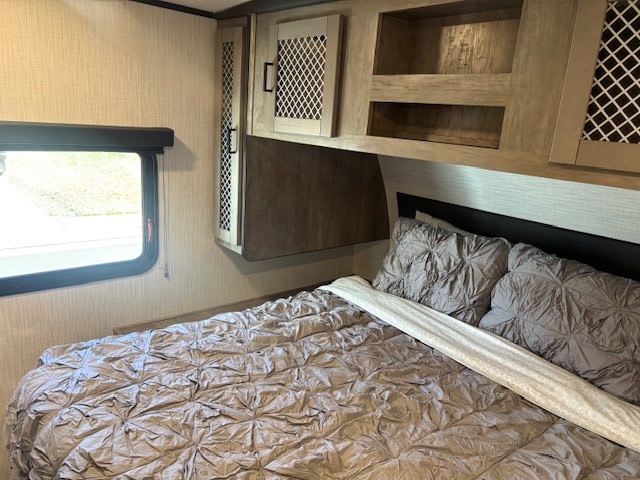 Cozy RV rental bedroom featuring a bed with gray comforter, pillows, window, and light wood overhead cabinets.. Heartland RVs North Trail 2021
