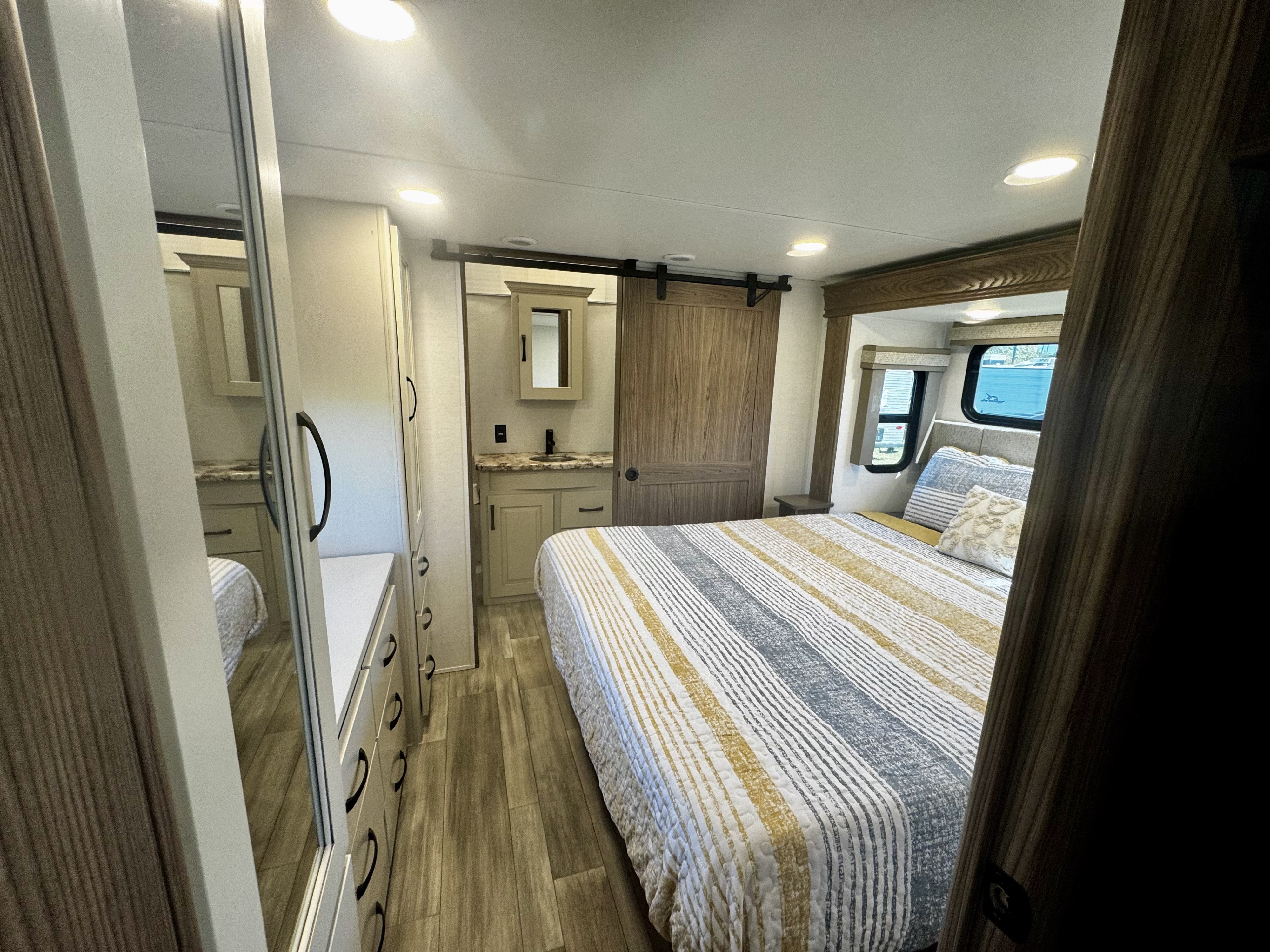Bright RV rental bedroom with striped bed, wood floors, full vanity, and barn door entry, promising comfortable stays.. Alliance RV Paradigm 2024