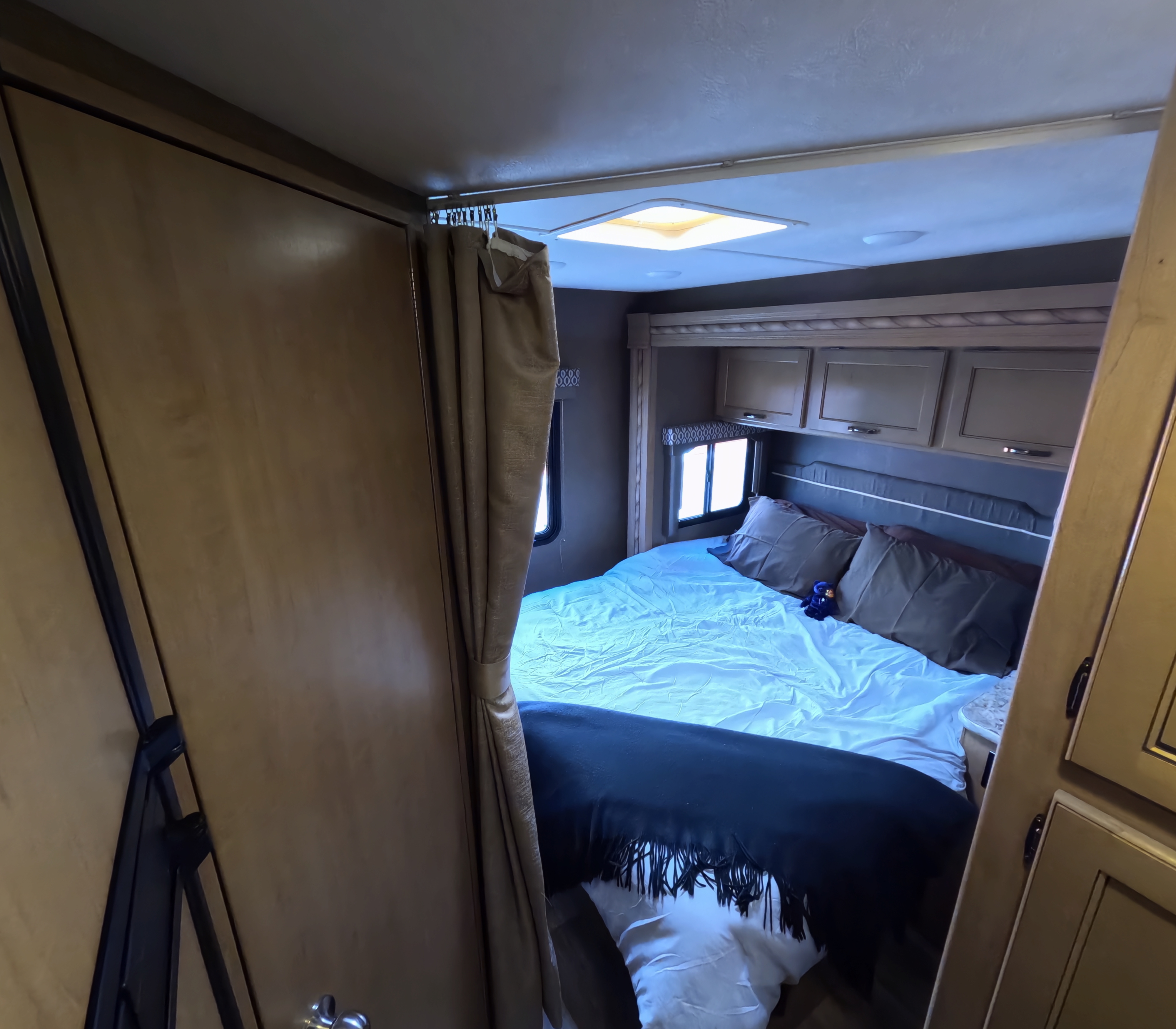 This RV rental bedroom features a cozy bed with light blue sheets and a black blanket, overhead cabinets, and windows.. Thor Freedom Elite 2018