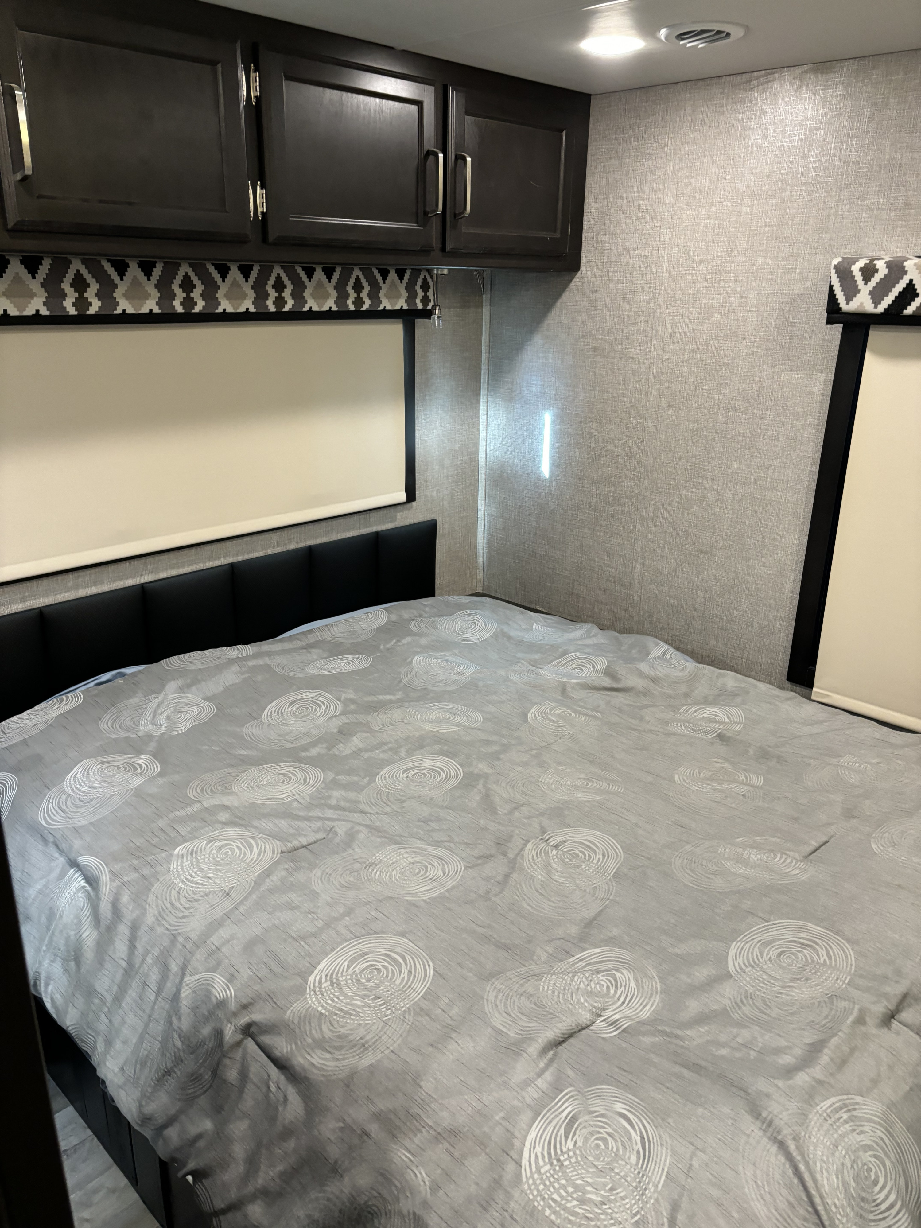 Cozy RV rental bedroom featuring a grey patterned comforter, black headboard, overhead cabinets, and textured walls.. Jayco Greyhawk 2022