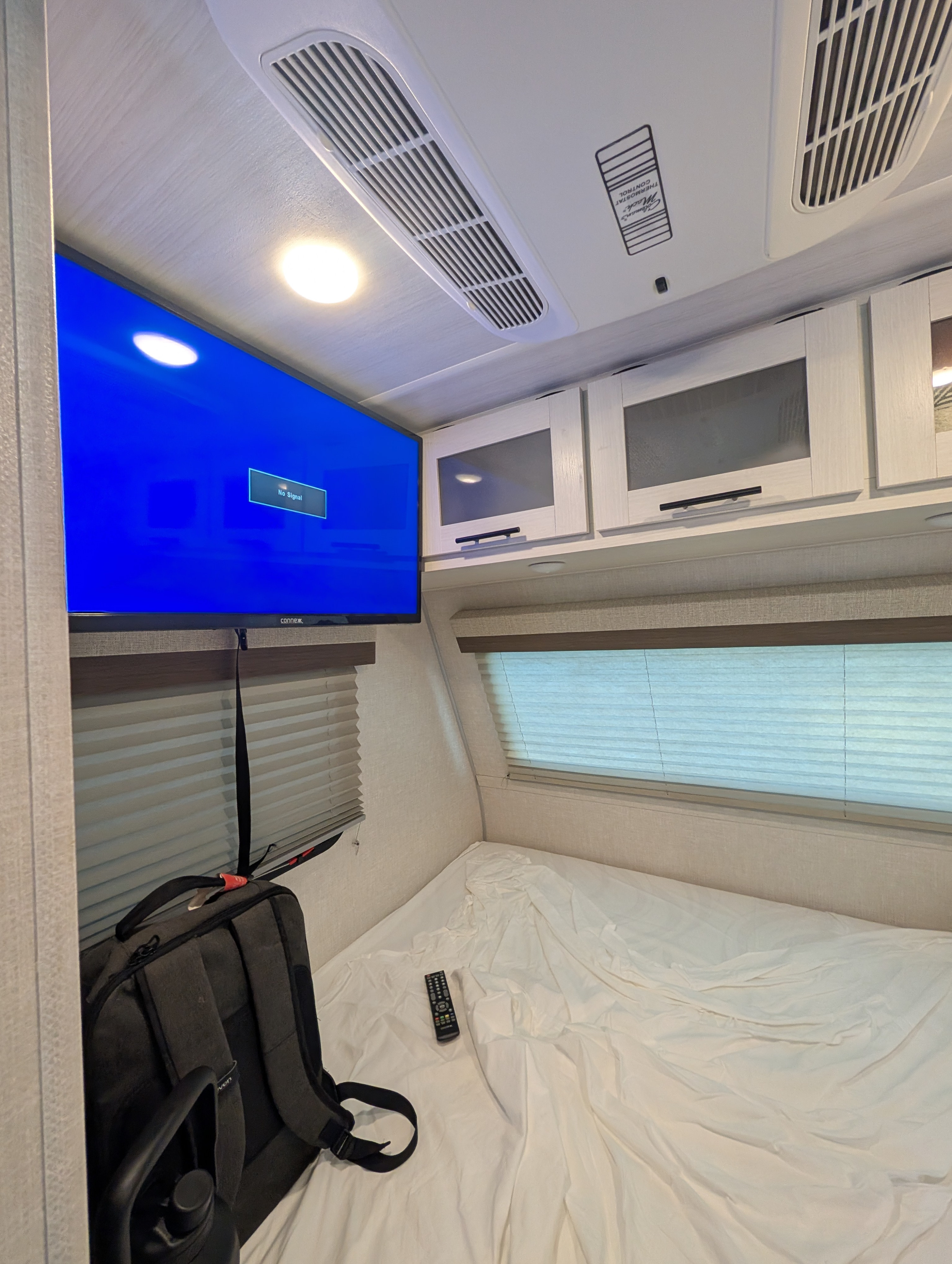 Inside an RV rental bedroom: A mounted TV, AC unit, overhead cabinets, and a bed with a remote and backpack.. No Boundaries 16 2024