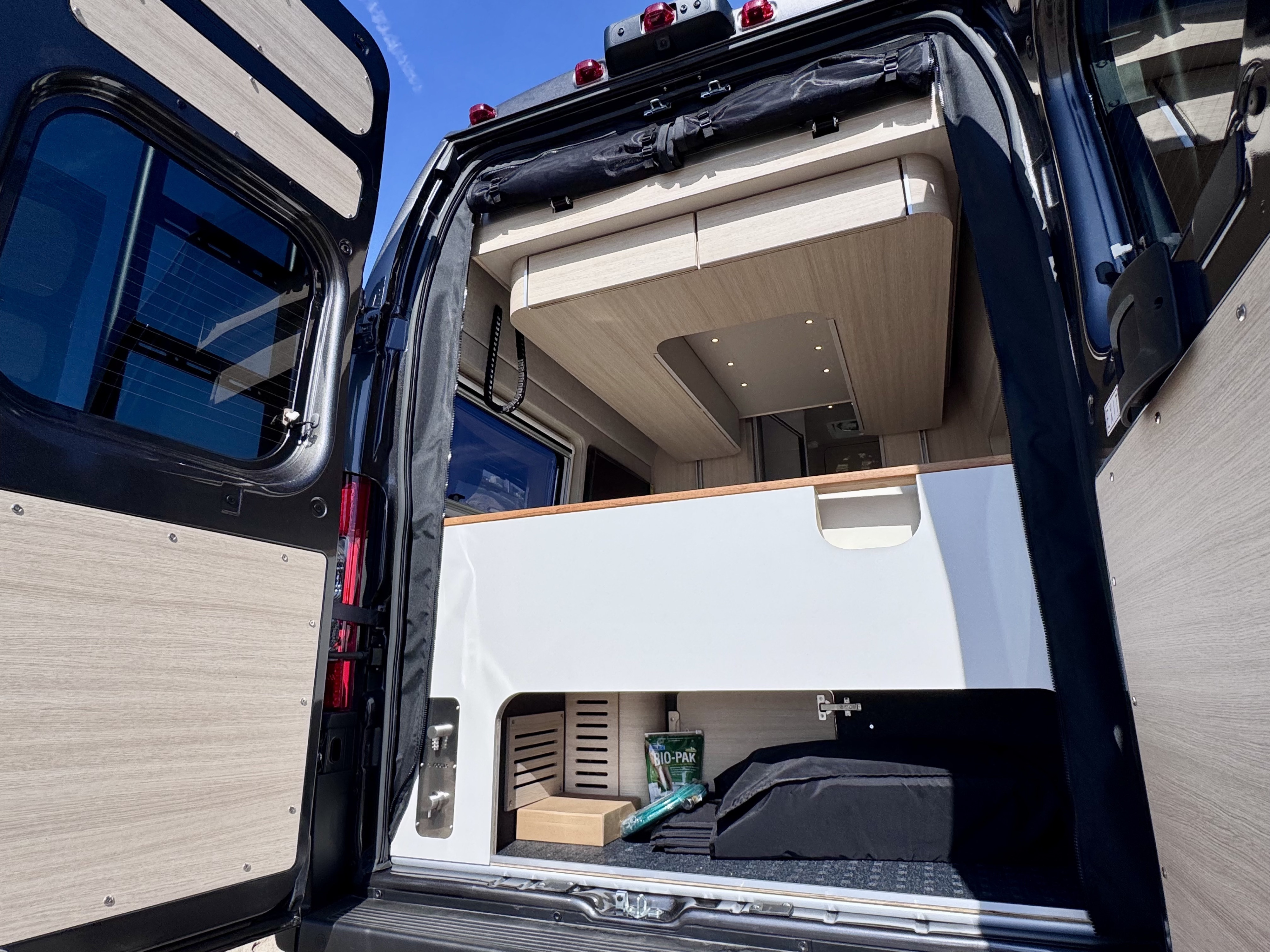 Open rear doors of an RV rental revealing its interior: a white bed/storage unit, lower storage with gear, wood ceiling, and lights.. Noovo Plus LB 2025