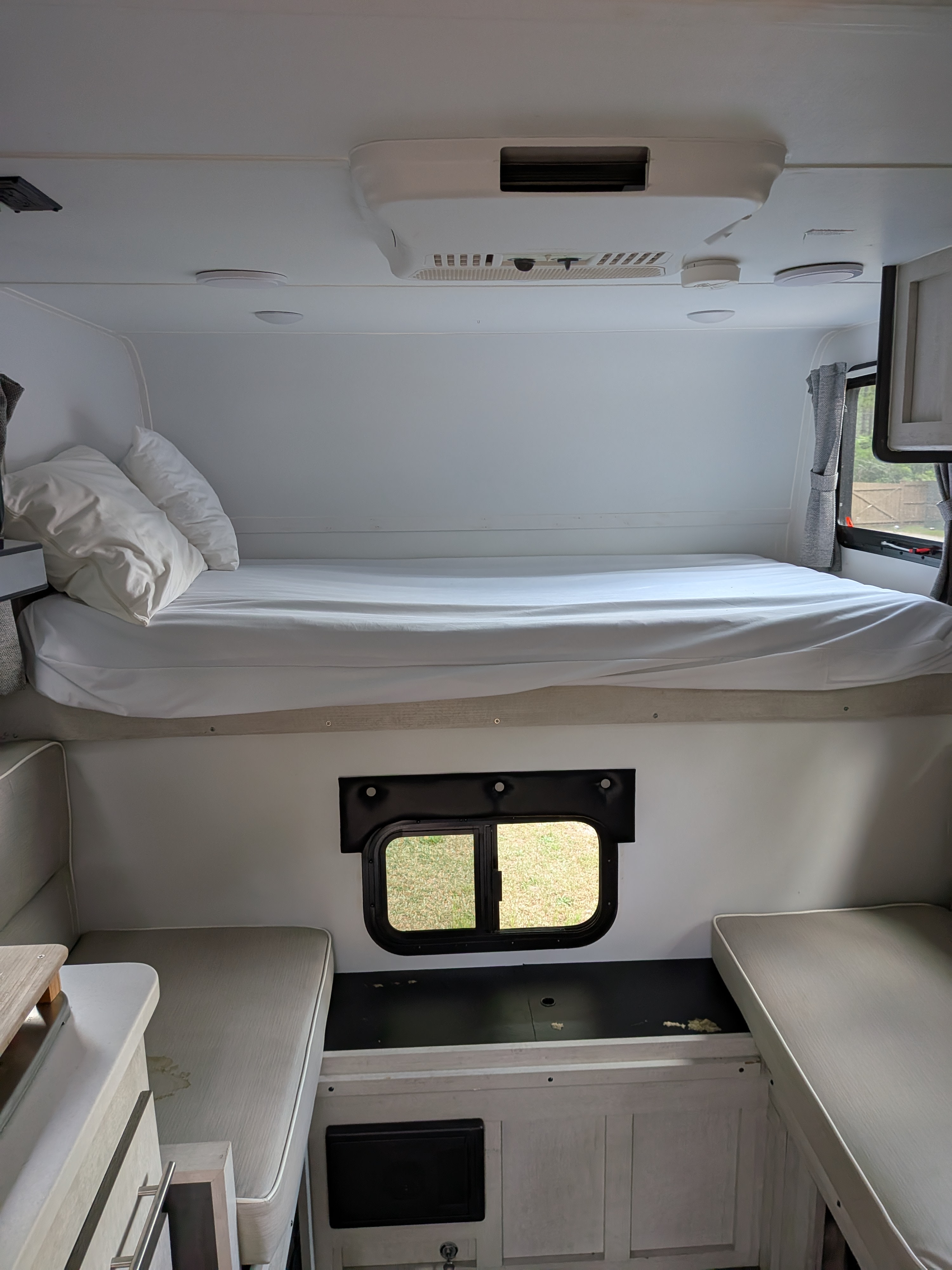 Cozy RV rental interior with a raised bed, white linens, ceiling AC, and lower dinette seating near a window.. Travel Lite 770sl 2022