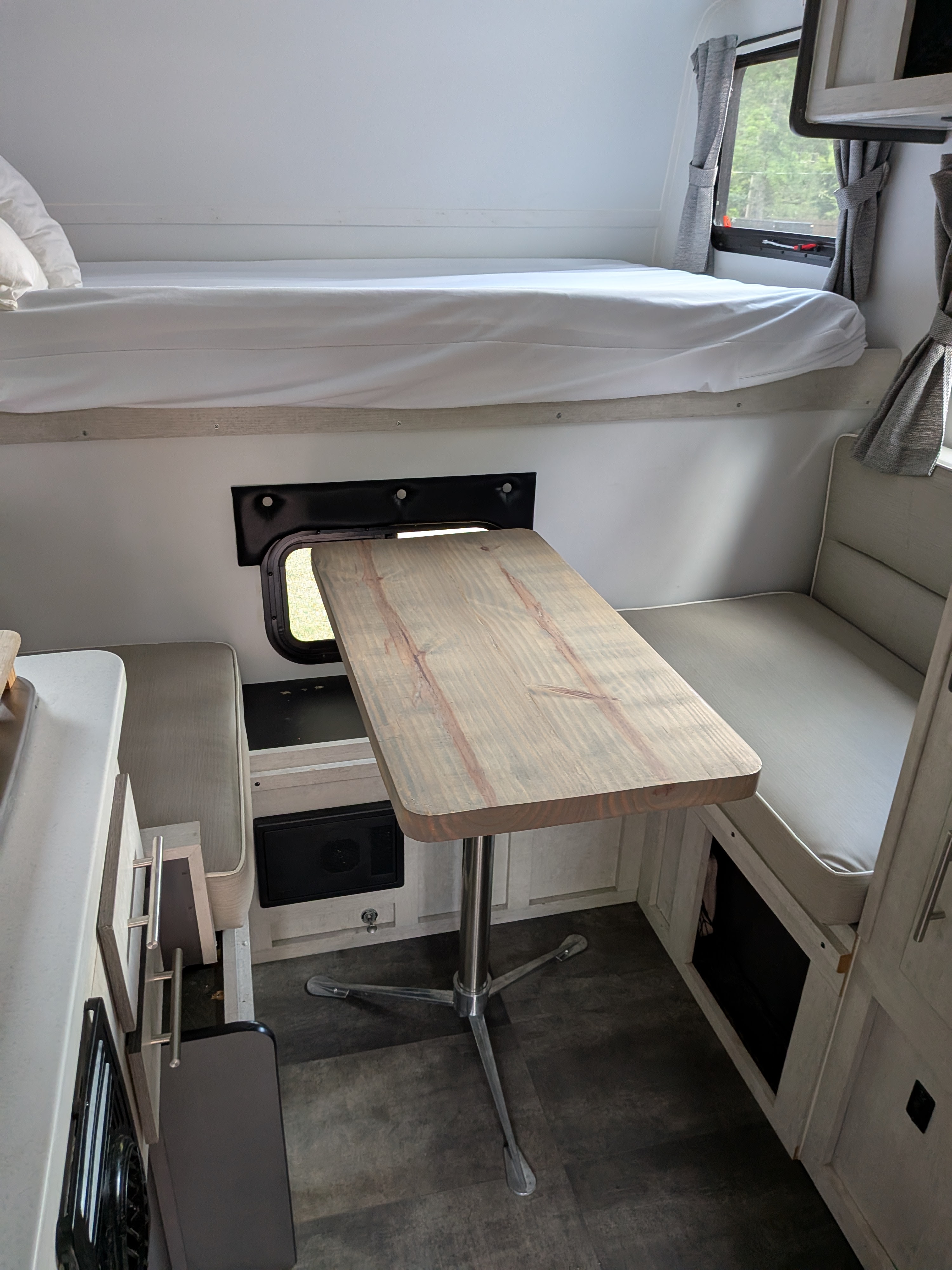 RV rental interior showing an overhead bunk with white sheets, a wooden table, and upholstered dinette seating.. Travel Lite 770sl 2022