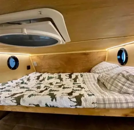 Inside a cozy RV rental: A bed with a moose-patterned comforter, plaid sheets, wooden walls, and small round windows.. Bend Teardrop Teardrop 6x12 2023