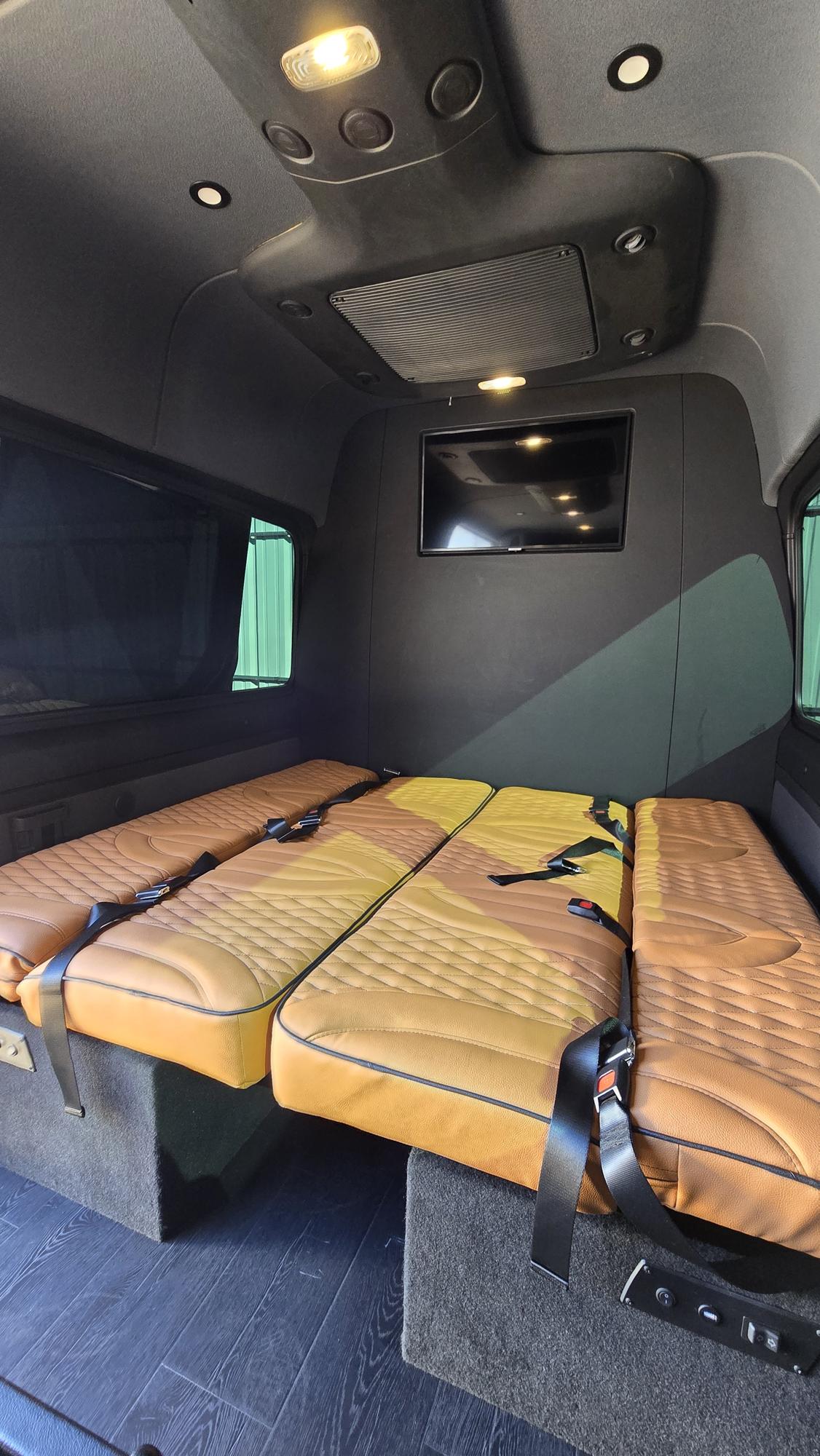 RV rental van interior with three tan quilted seats/beds, seatbelts, wall-mounted TV, and overhead lighting.. Mercedes-Benz Sprinter 2022