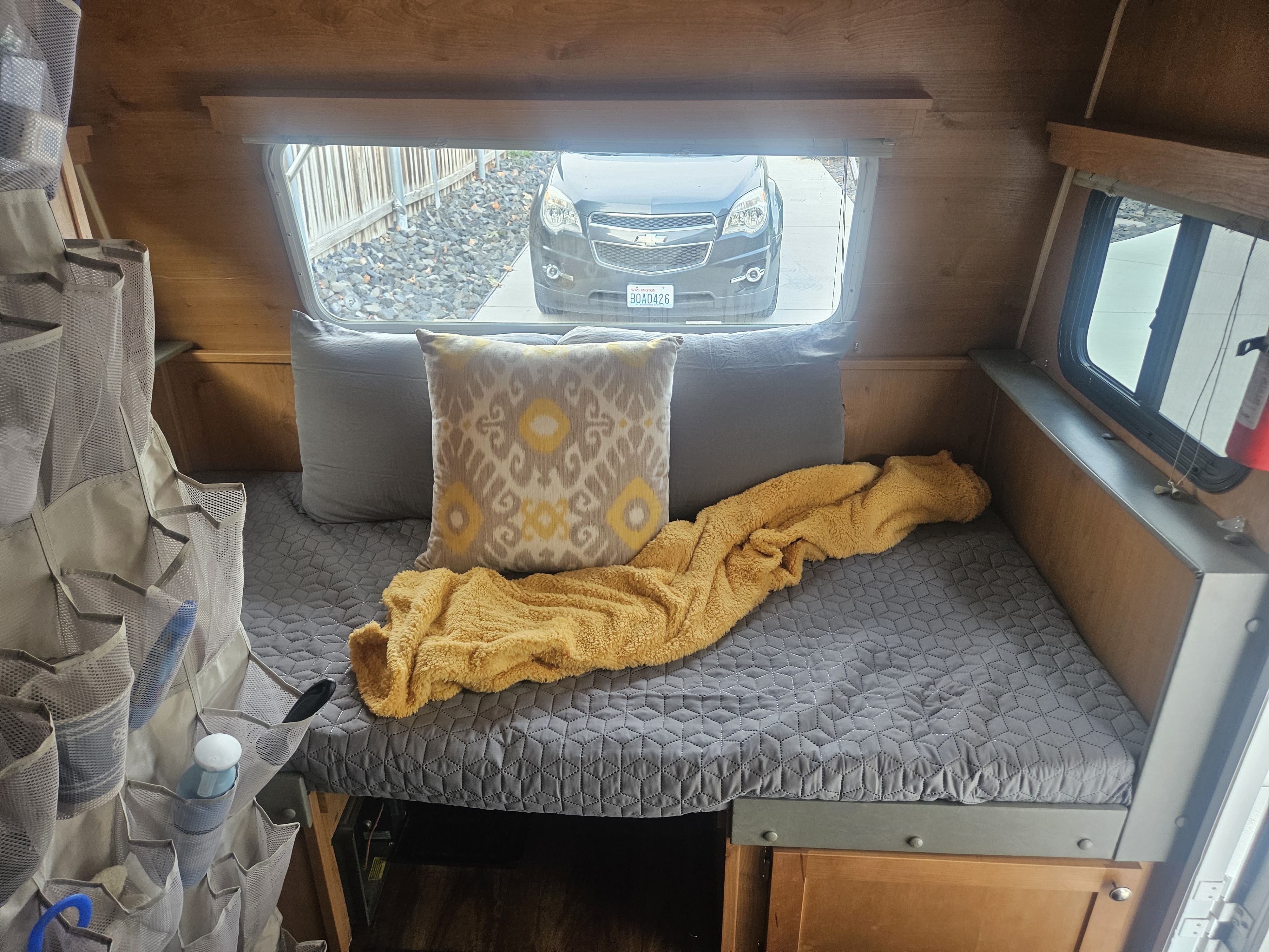 RV rental interior: Cozy bed with pillows & yellow blanket, window view of SUV, clever hanging storage.. Rivt Retro 166 2017