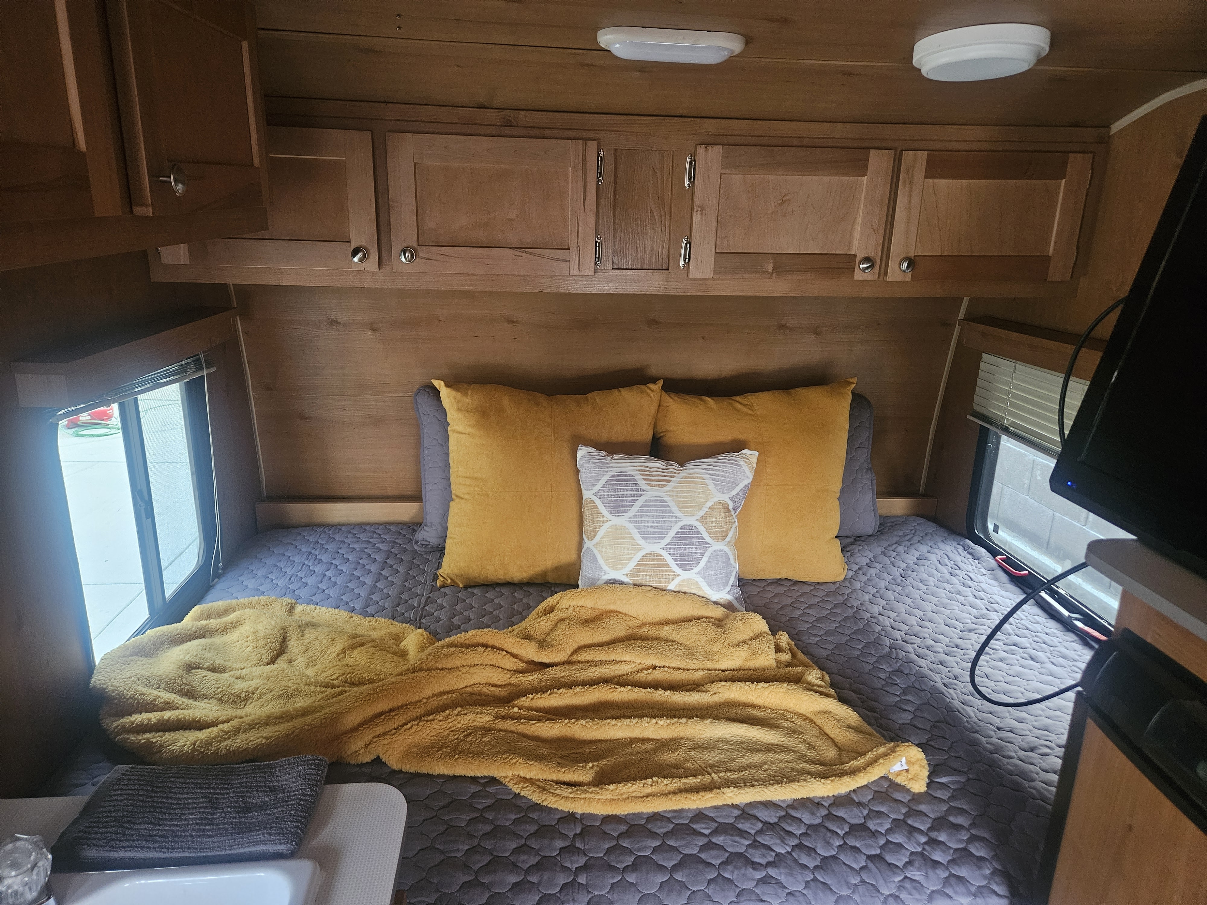Inside an RV rental: a cozy bed with a gray quilt, yellow pillows, and a fuzzy blanket under wooden cabinets.. Rivt Retro 166 2017