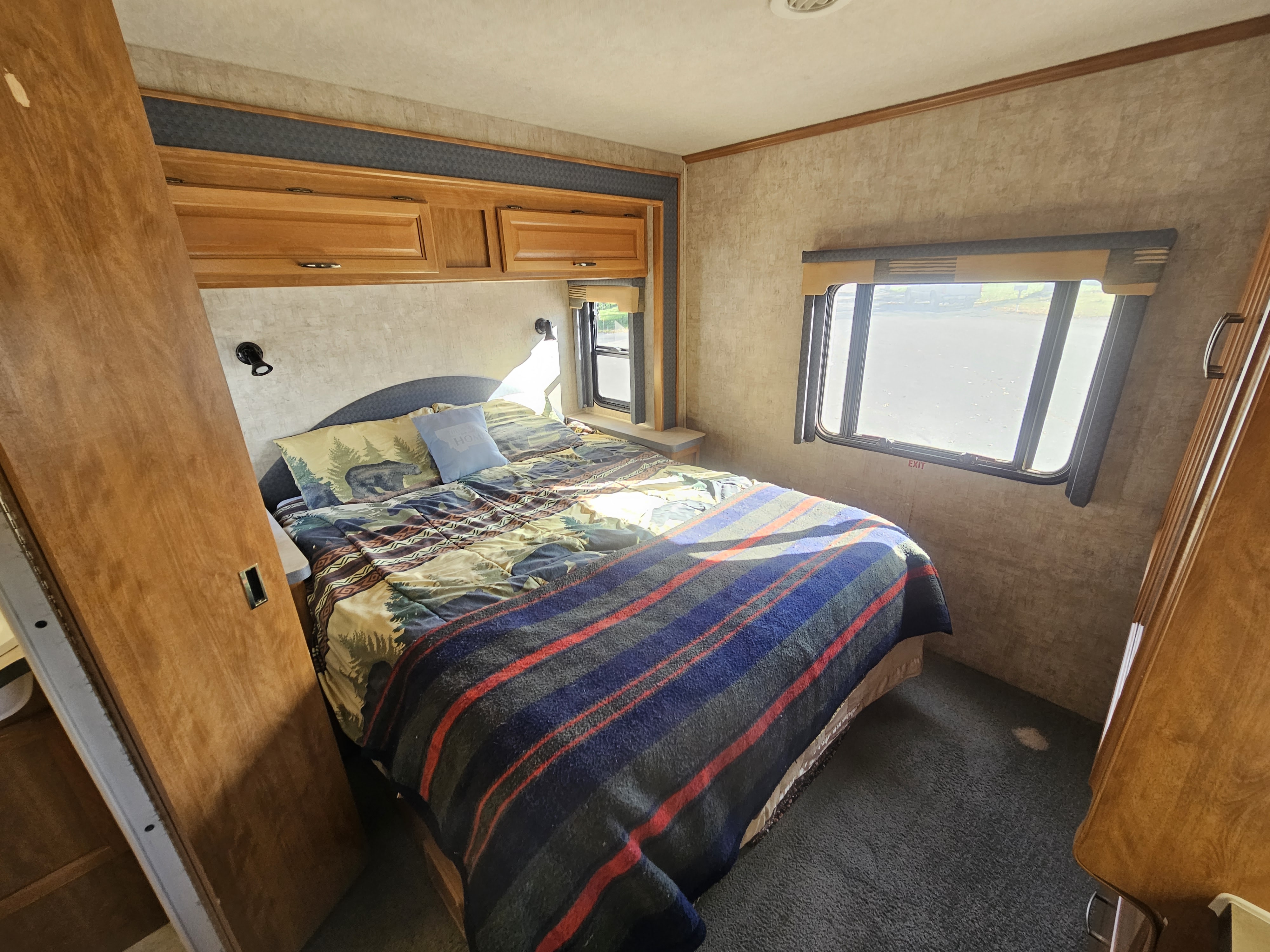 Comfortable RV rental bedroom featuring a queen bed with nature-themed bedding, overhead cabinets, and bright windows.. Tioga SL 2006