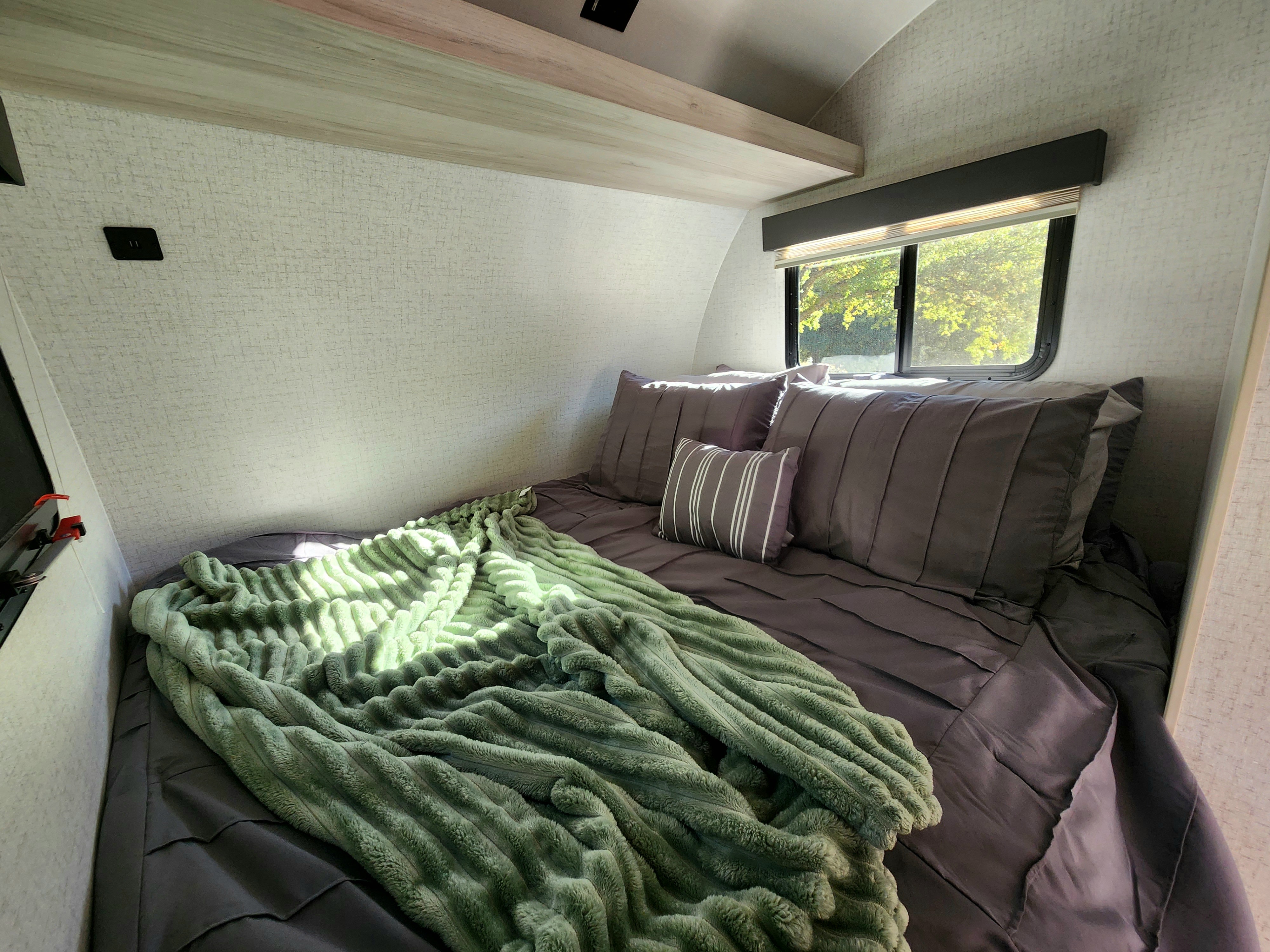 Inside an RV rental: a snug bedroom with a grey bed, plush green blanket, and bright window for relaxing.. Winnebago Hike 2021