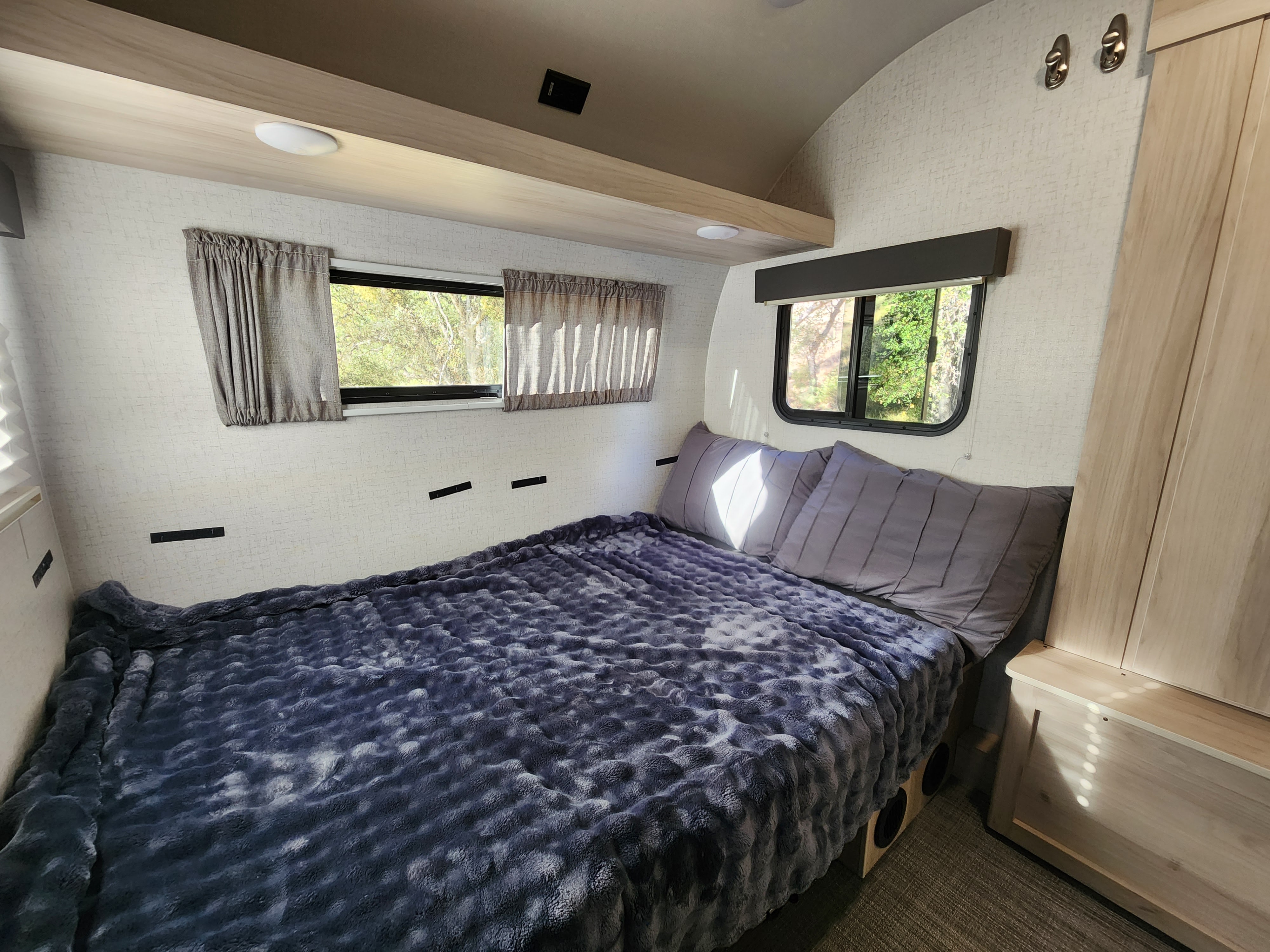 Inside this cozy RV rental bedroom, a plush bed with a textured blanket, windows showing nature views, and overhead storage.. Winnebago Hike 2021