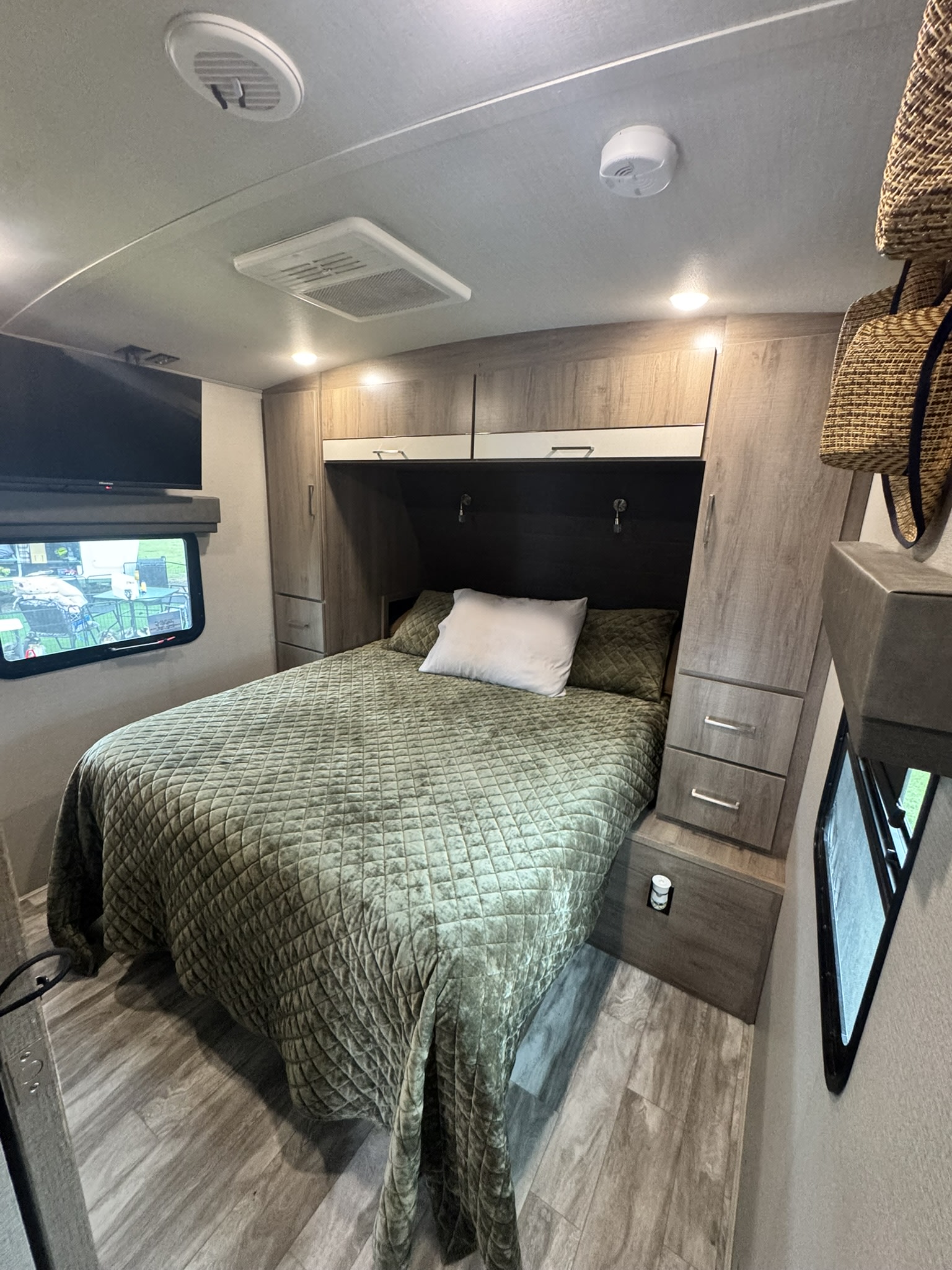 This RV rental bedroom features a cozy bed with green quilted bedding, wood cabinetry, a TV, and windows.. Grand Design Imagine 2023