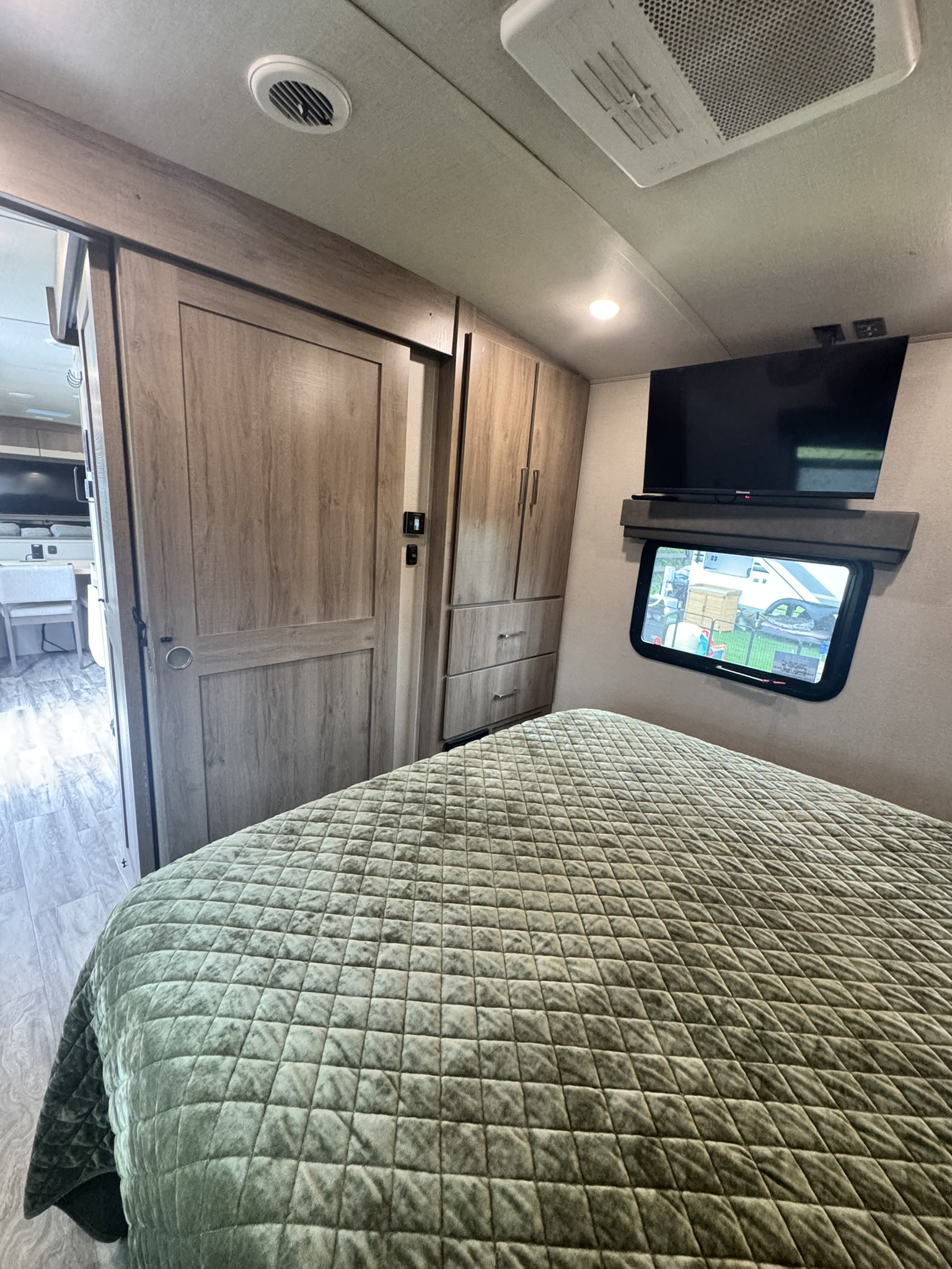 Cozy RV rental bedroom featuring a quilted bed, wall-mounted TV, window view, and wooden cabinetry. Ideal for your next adventure.. Grand Design Imagine 2023