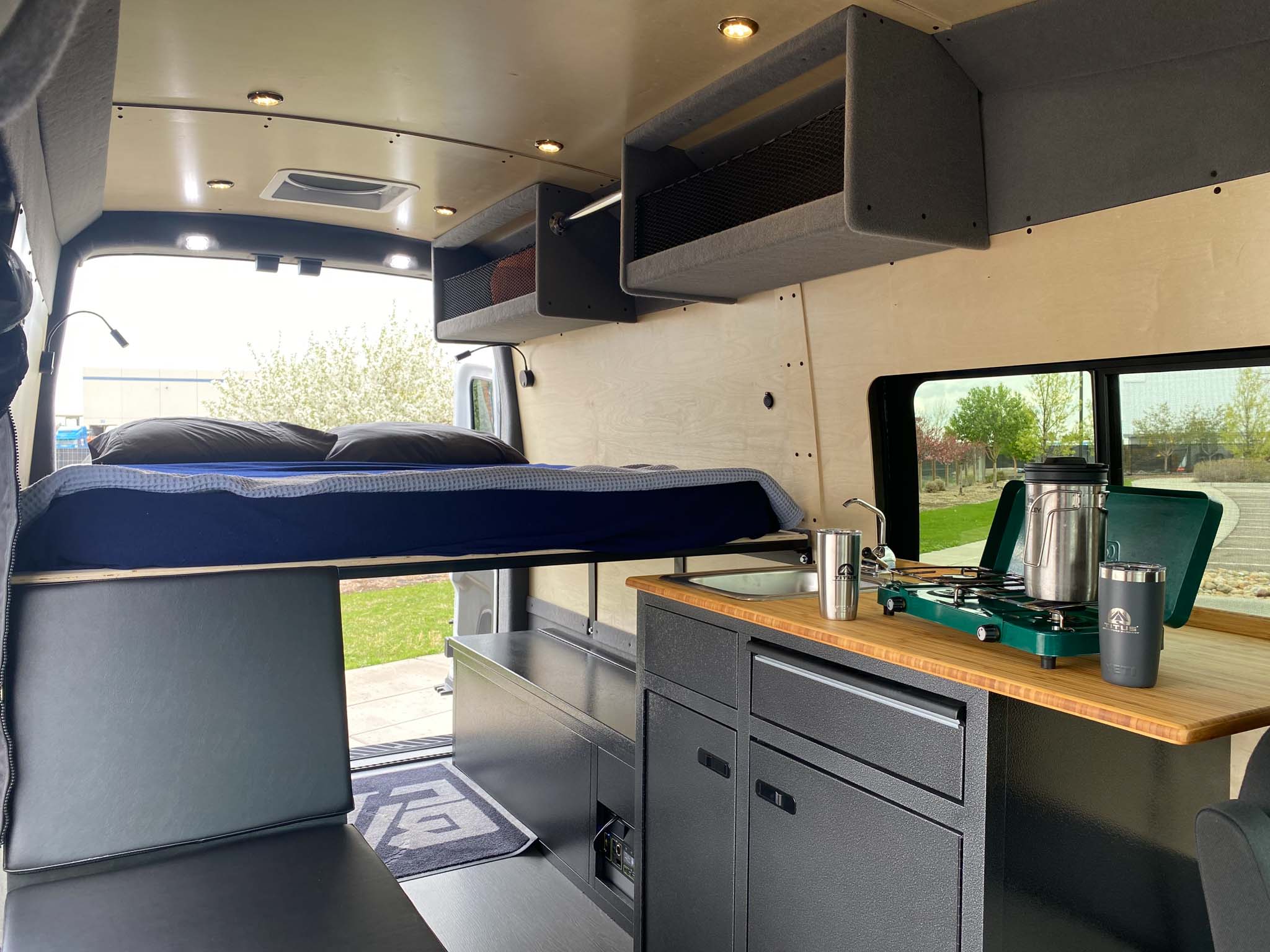 This RV rental interior offers a raised bed, functional kitchenette with stove & sink, and ample storage for van life.. Ford Transit 2023