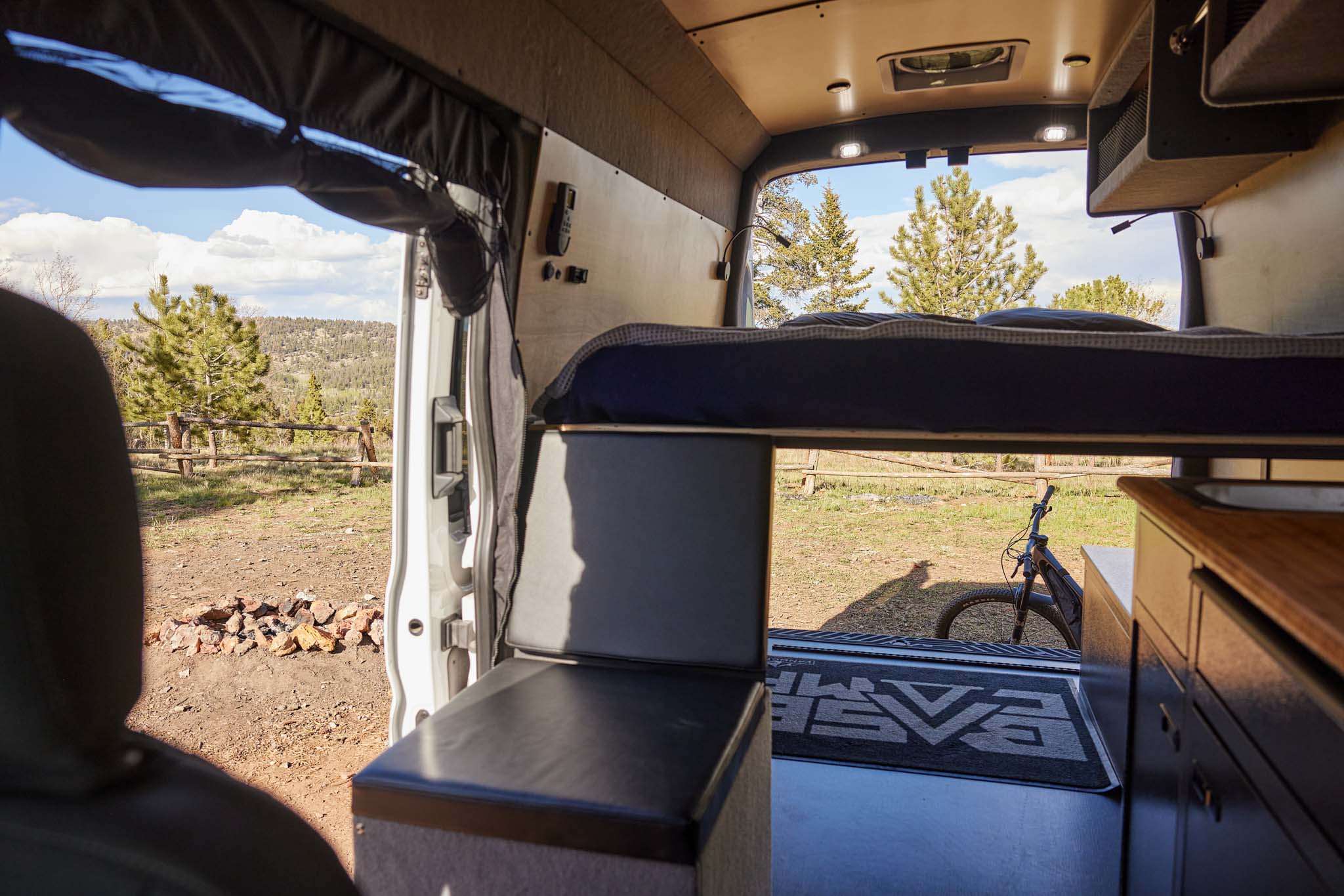 Inside an rv rental: elevated bed, bench, and kitchenette with a view of trees, mountains, and a bike out the open back.. Ford Transit 2023