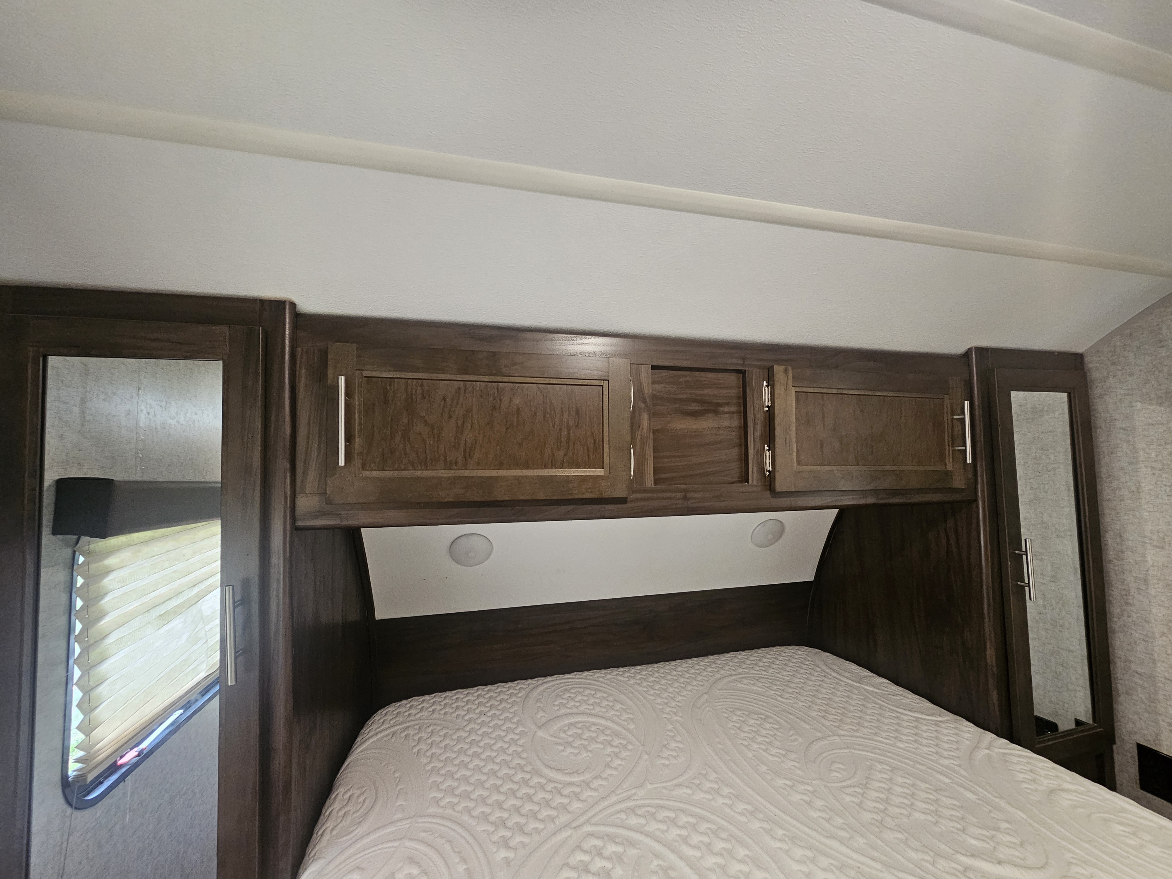 Inside this RV rental bedroom, observe a bed with patterned mattress, dark wood overhead cabinets, and mirrored side storage.. Forest River Cherokee 2018