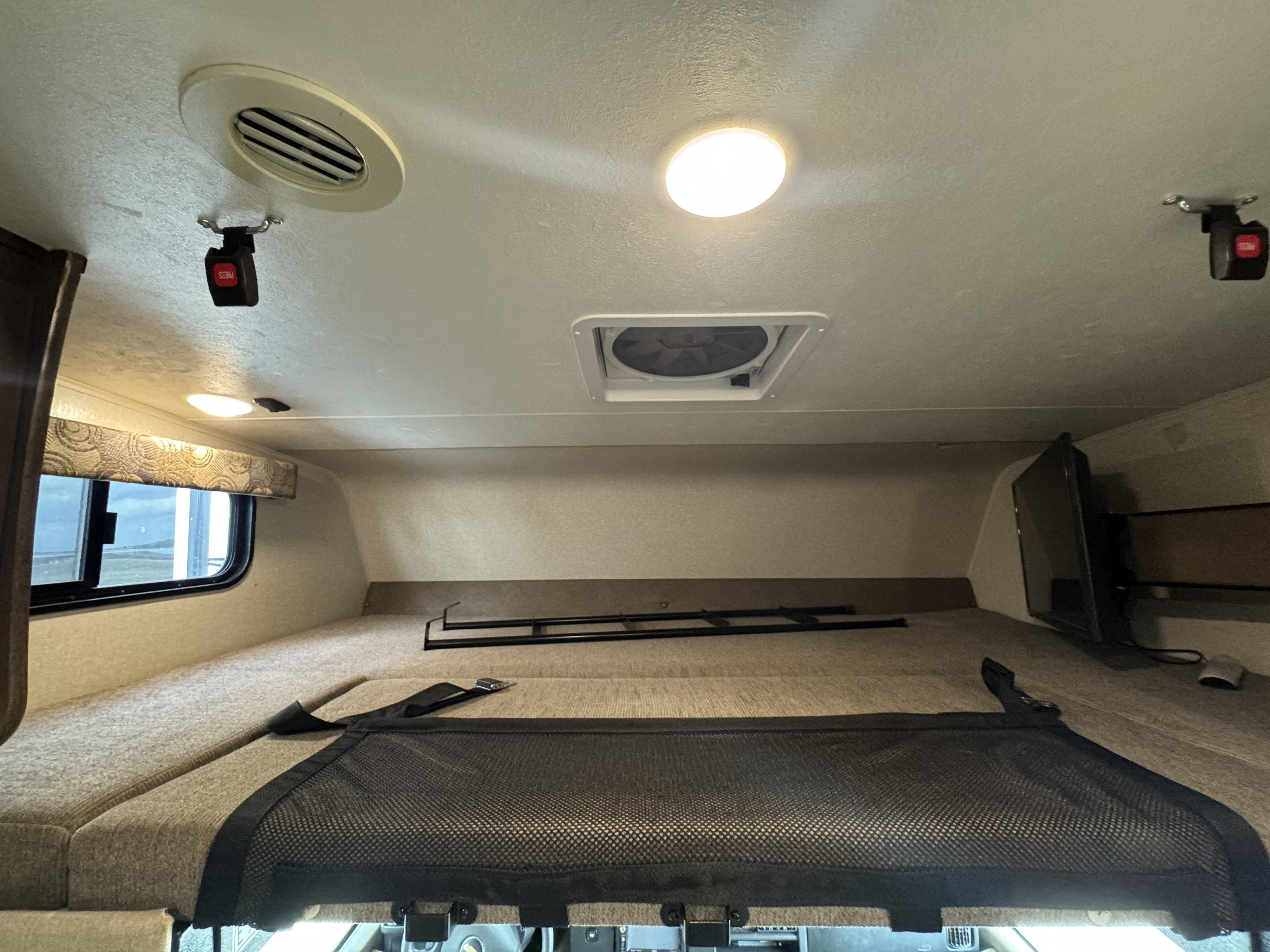 Overhead bunk in an RV rental, featuring a safety net, window, TV, AC vent, and ceiling fan for a comfortable stay.. Forest River Coachmen Freelander 2019