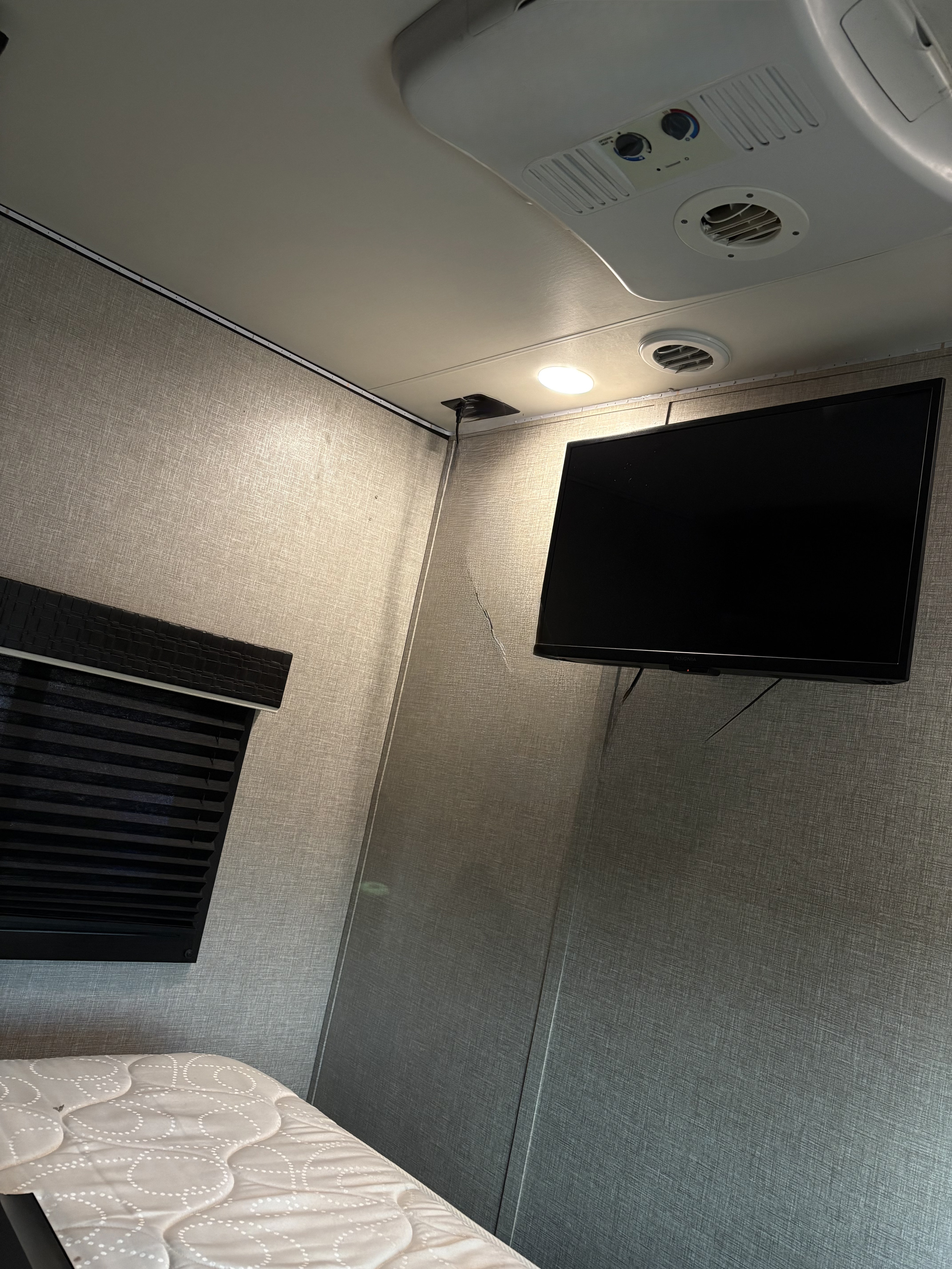 RV rental interior featuring a bed, mounted TV, AC unit, window blind, and textured walls. Visible wires and a small wall crack.. Jayco Jay Flight 2022