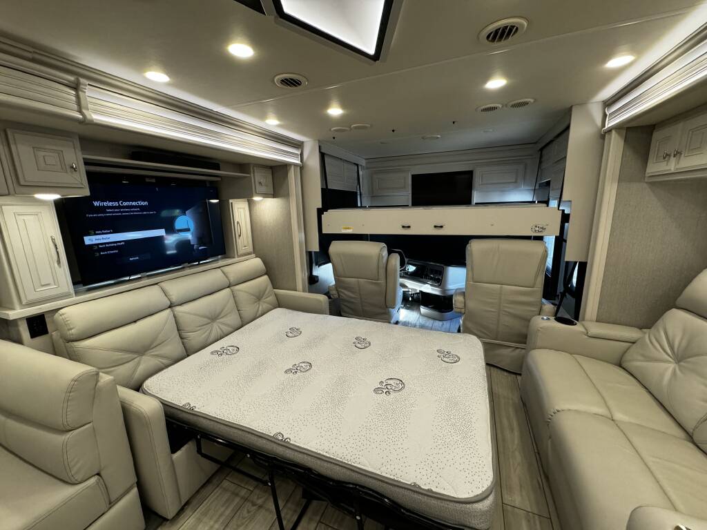 Inside a modern RV rental, showing a comfy pull-out bed, large TV, cream sofas, and the driver's area beyond.. Holiday Rambler Armada 2022