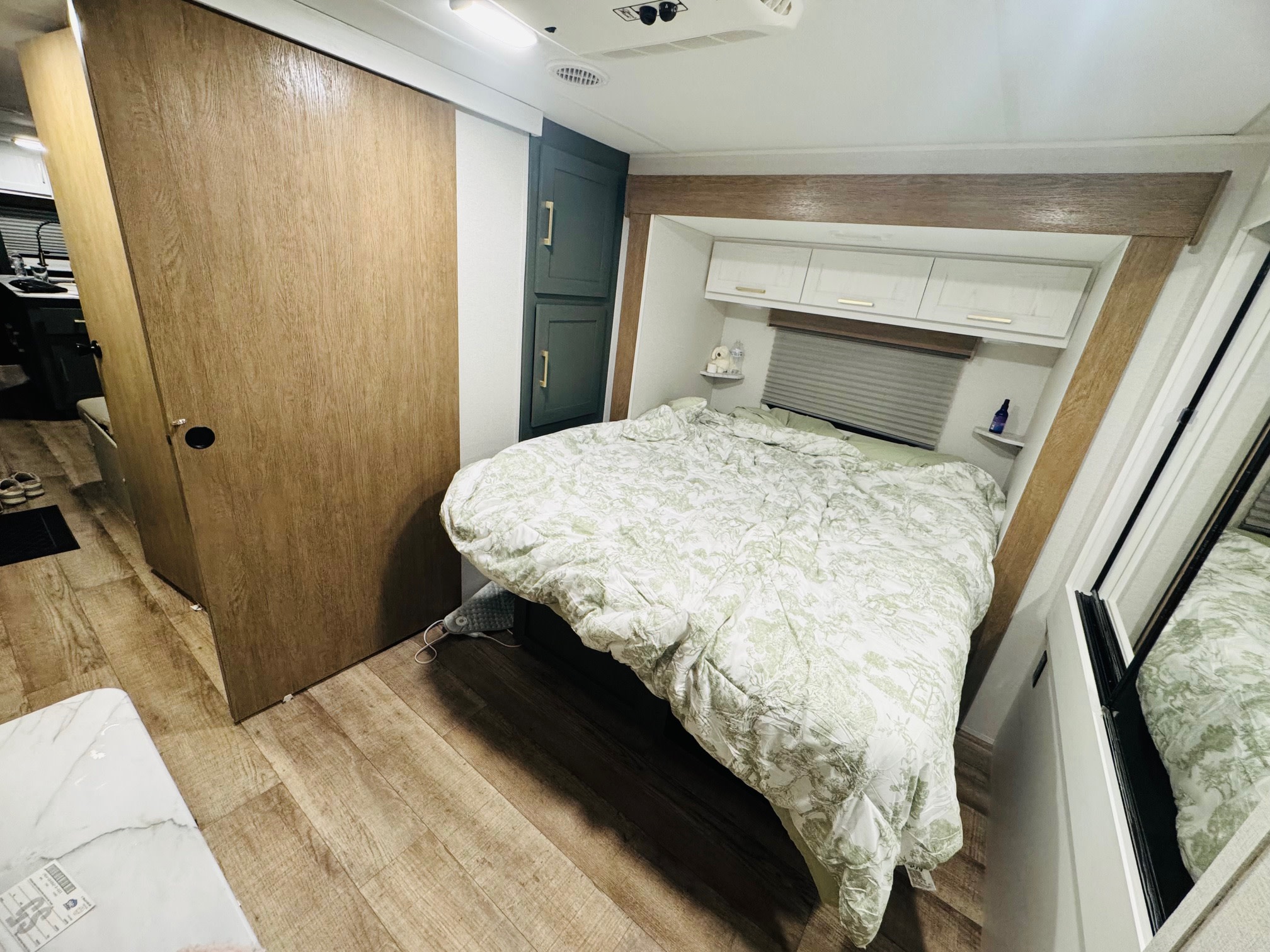 This RV rental interior shows a cozy bedroom with green bedding, wood accents, overhead cabinets, and a sliding door.. Heartland RVs Eddie Bauer 2025