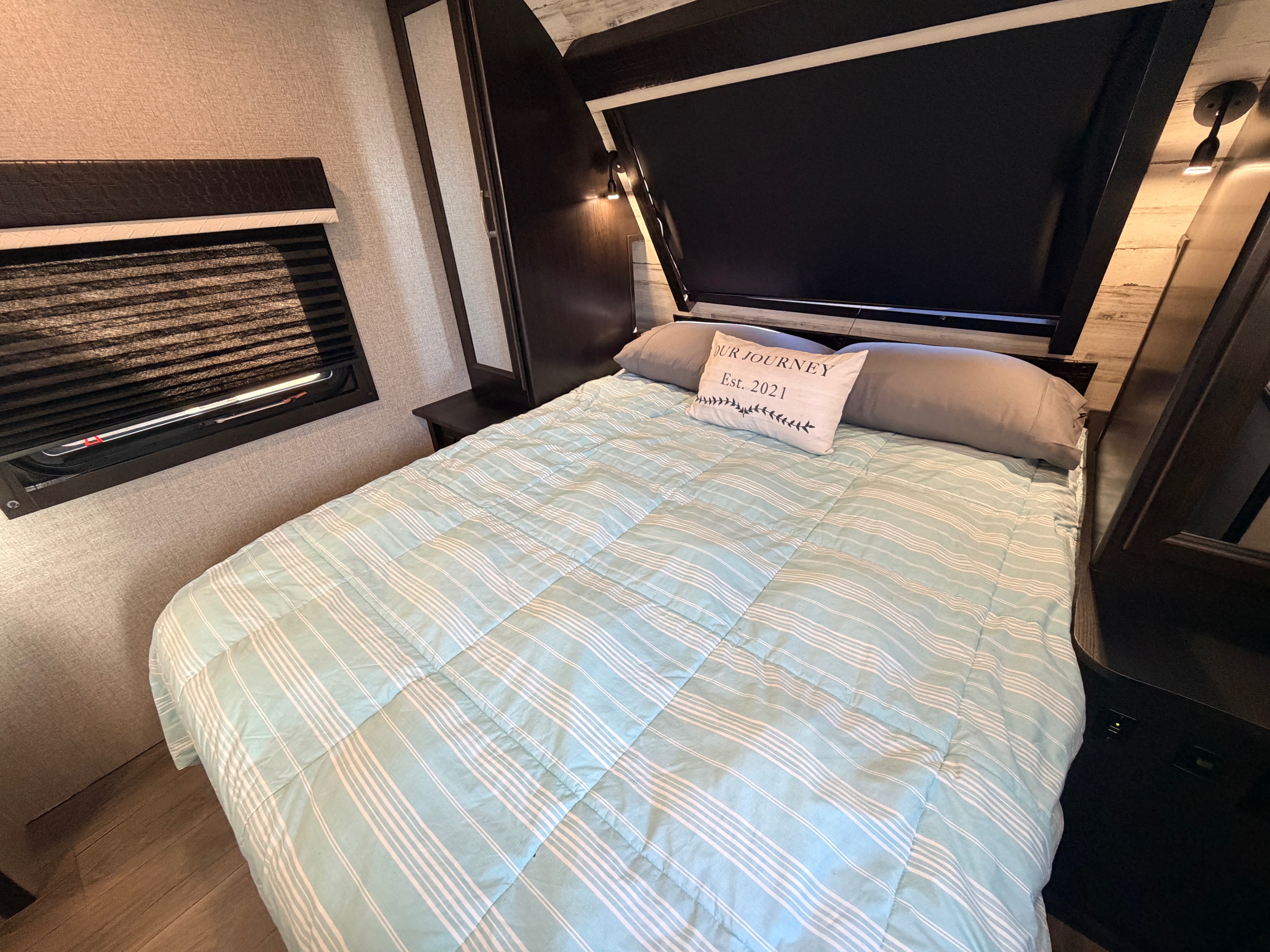 Inviting RV rental bedroom with striped blue bedding, dark wood decor, window blinds, and a "Our Journey" pillow.. Jayco White Hawk 2021