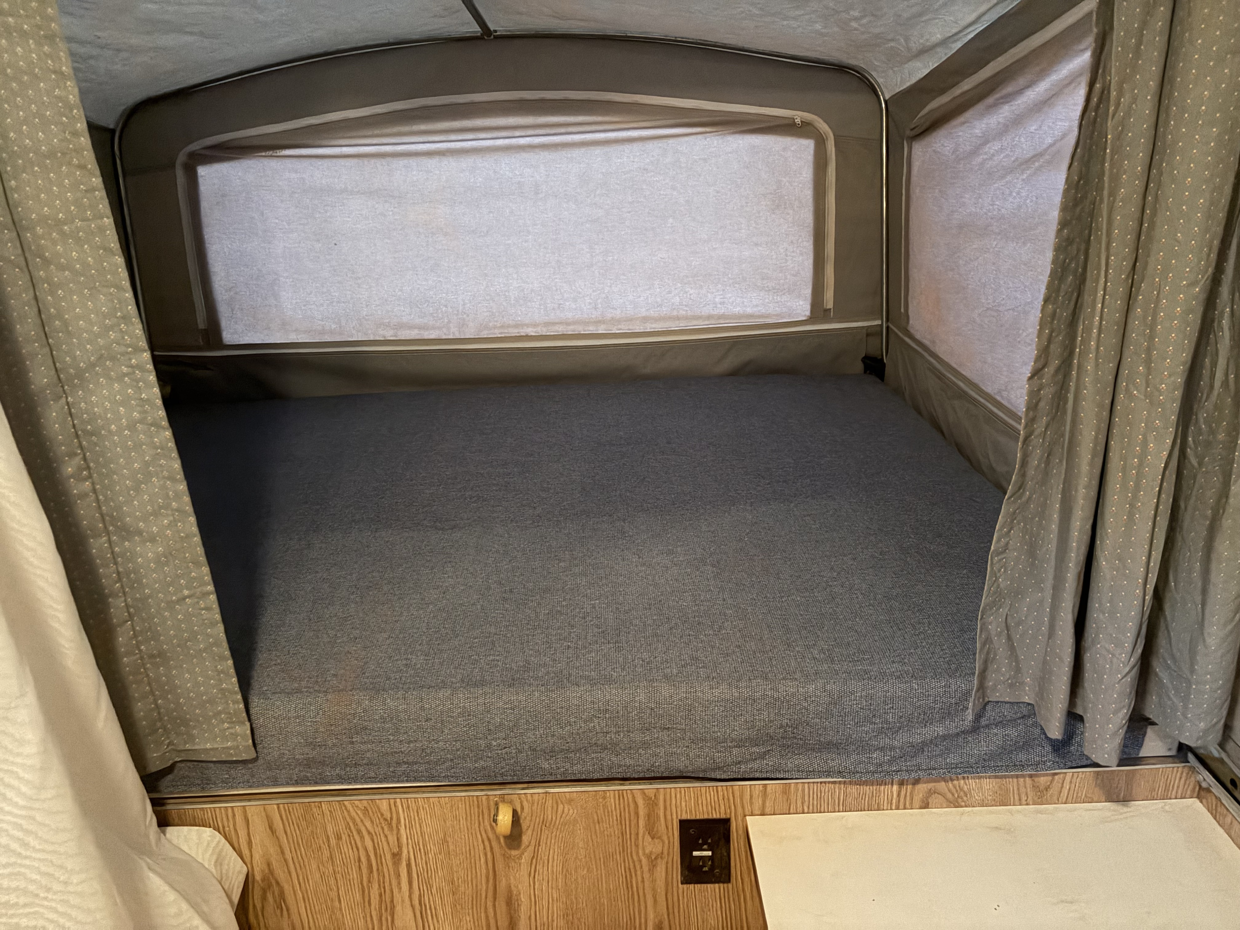 Cozy bed area in an RV rental, featuring a gray mattress, window, and patterned curtains. Perfect for camping trips.. Dutchmen M1204 1996