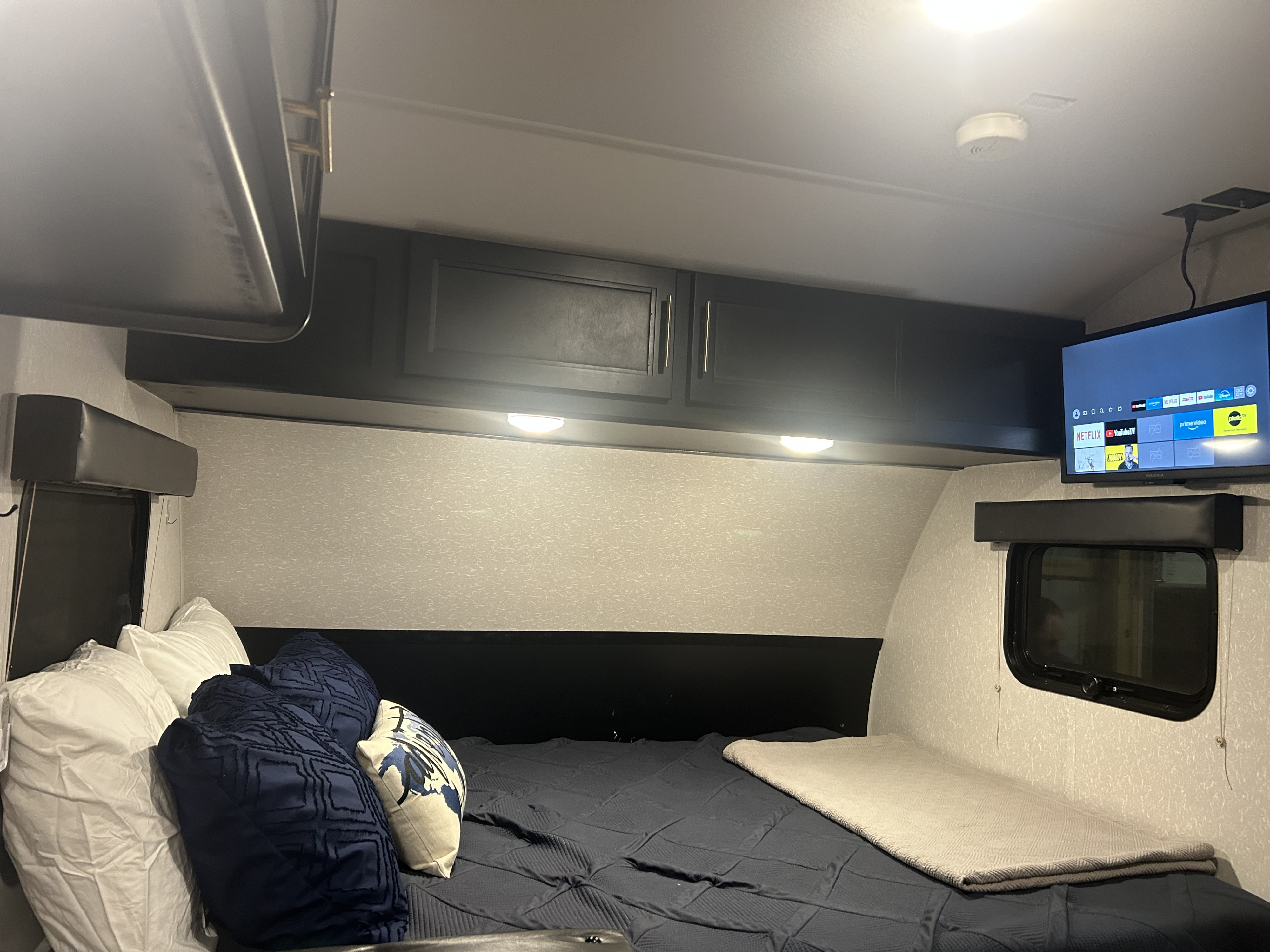 Cozy RV rental bedroom featuring a large bed with navy bedding, dark overhead cabinets, accent lights, and a wall-mounted TV.. Forest River Cherokee Cherokee Black Label 2021
