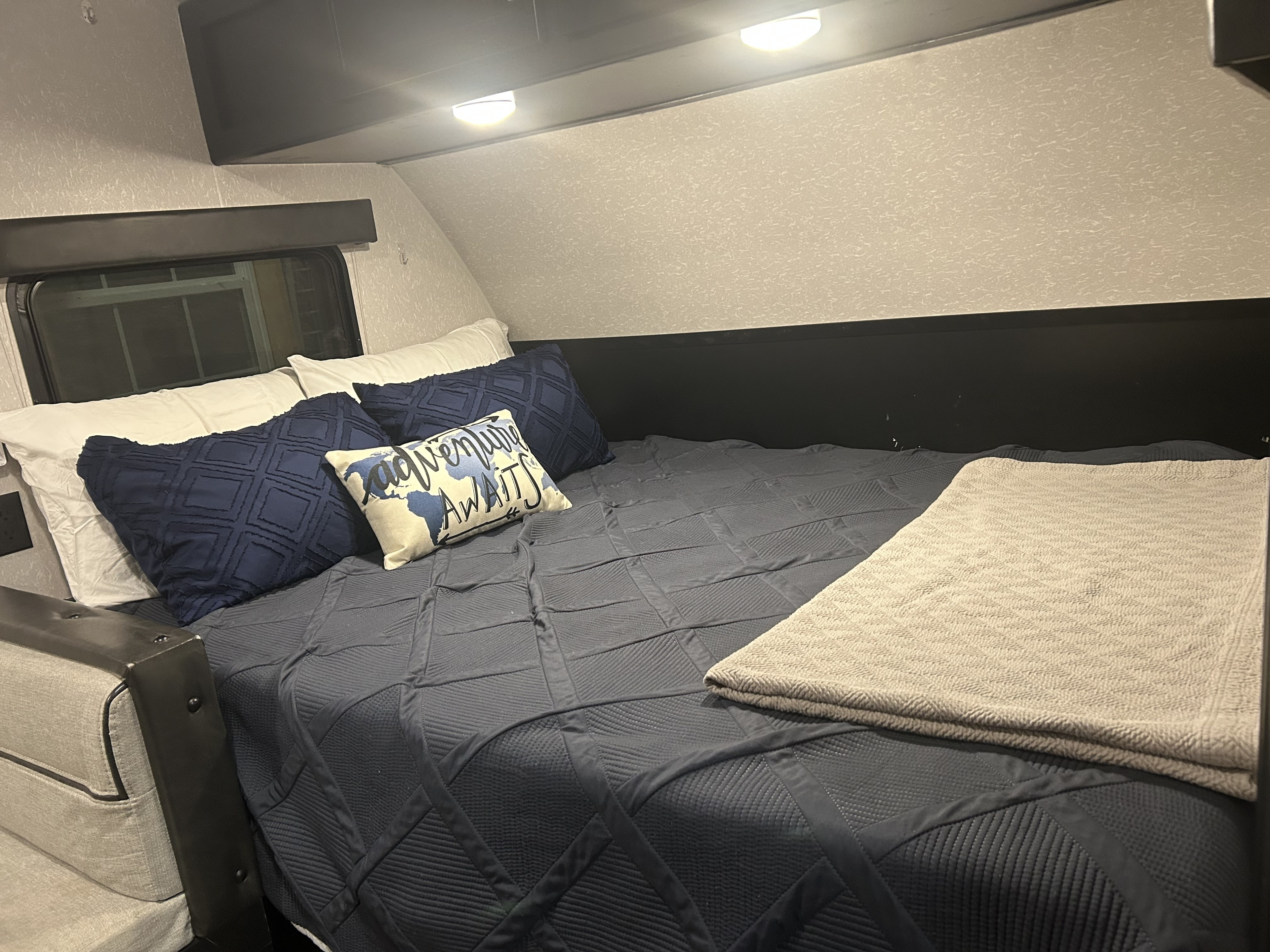 Cozy RV rental bed area with quilted blue bedding, accent pillows including an 'Adventure Awaits' cushion, and soft lighting.. Forest River Cherokee Cherokee Black Label 2021