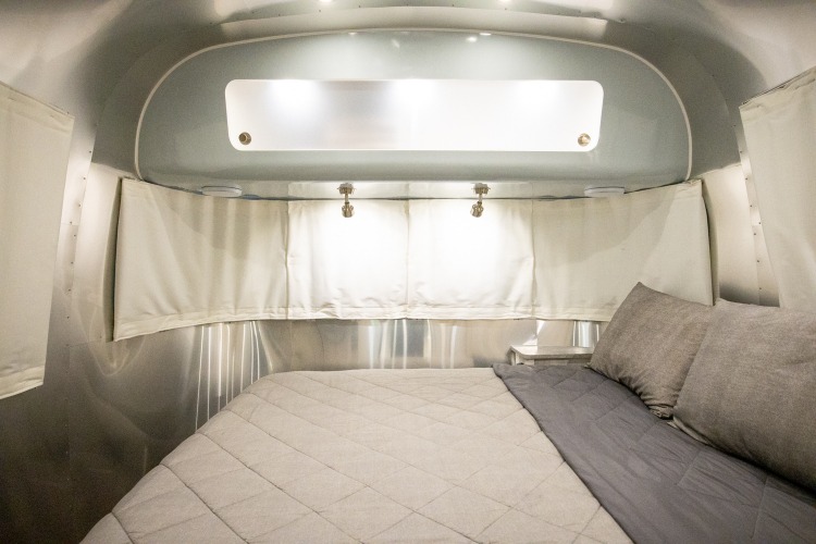 Comfortable RV rental bedroom features a cozy bed with gray pillows, white window shades, and modern lighting in a sleek aluminum interior.. Airstream International 2021