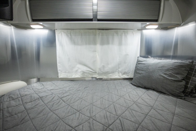 RV rental bedroom with a grey quilted bed, aluminum walls, white fabric curtains, and overhead lights.. Airstream Sport 2019