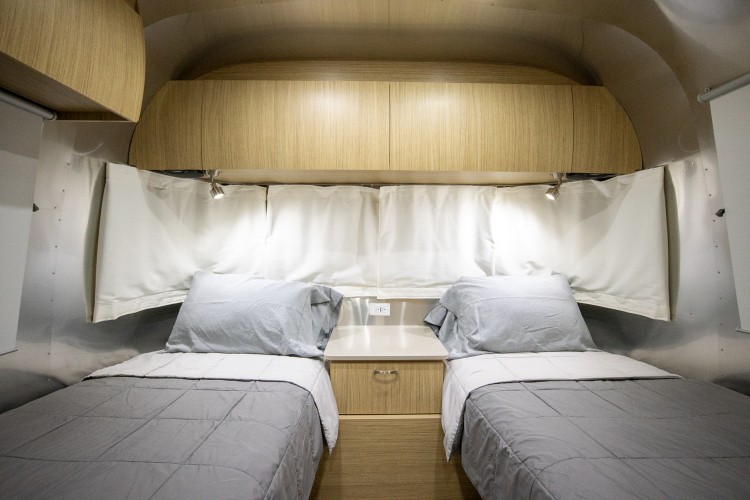 Comfortable twin beds in an RV rental bedroom, featuring overhead storage, a nightstand, and reading lights for a cozy stay.. Airstream Flying Cloud 2019