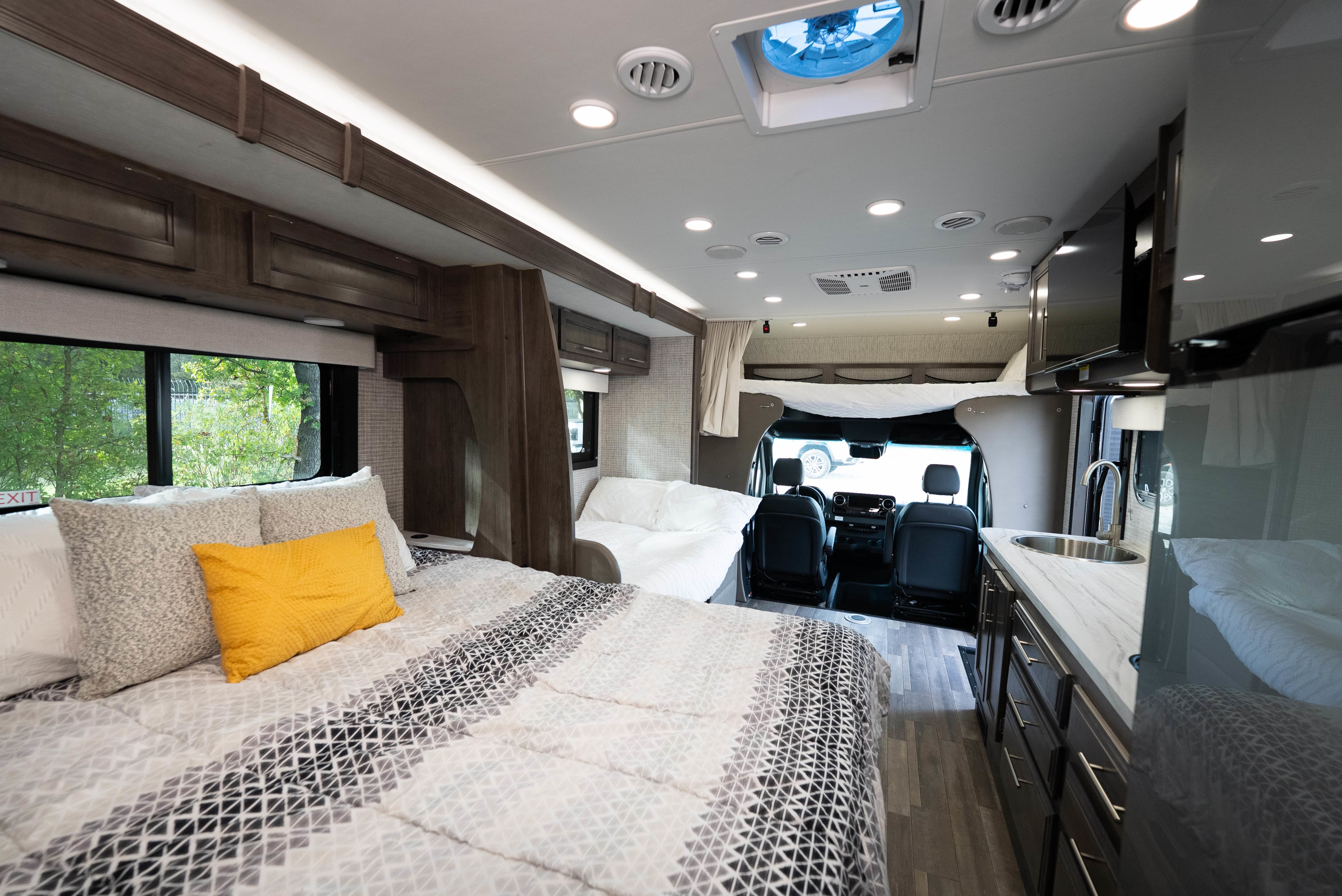 This rv rental interior features a cozy bedroom with patterned bedding, a compact kitchen with a sink, and the front cockpit.. Entegra Qwest 2025