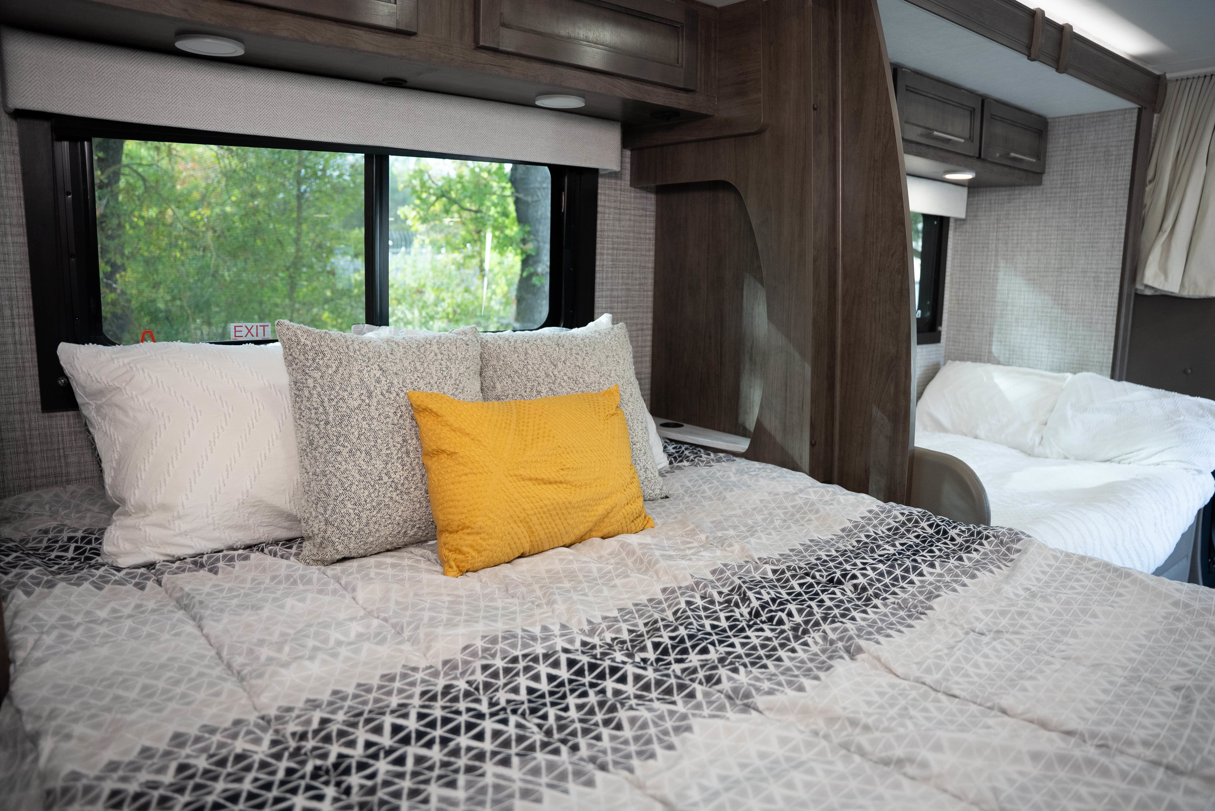Cozy RV rental bedroom with a bed featuring a geometric comforter, white, grey, and yellow pillows, and a scenic window view.. Entegra Qwest 2025