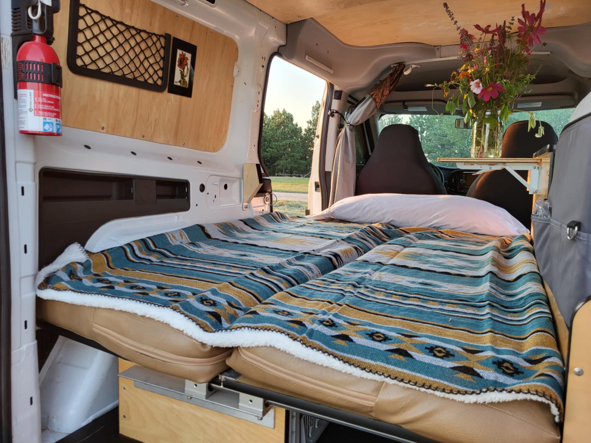 Cozy RV rental interior featuring a comfortable bed, wooden paneling, a fire extinguisher, and a nature view. Ideal for van life.. Dodge Dodge Promaster 2019