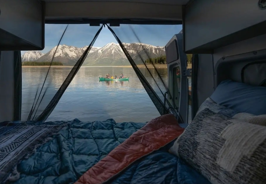A cozy RV rental bed with a stunning view of a lake, snow-capped mountains, and a canoe carrying people.. Winnebago Solis 2025