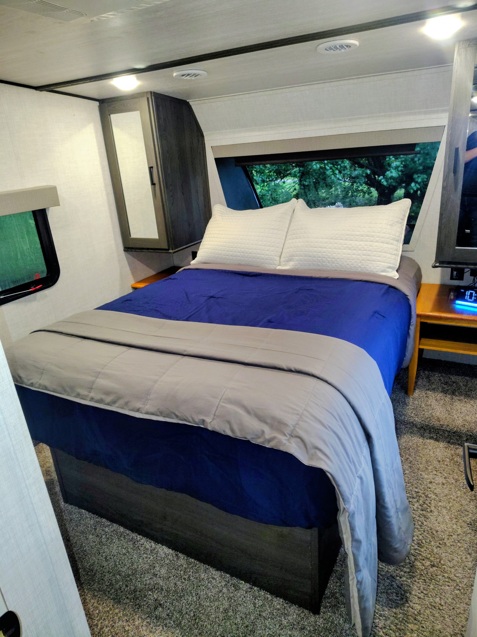 Explore this comfy RV rental bedroom: blue sheets, grey comforter, nightstand with clock, and windows with nature views.. Forest River Impression 2023